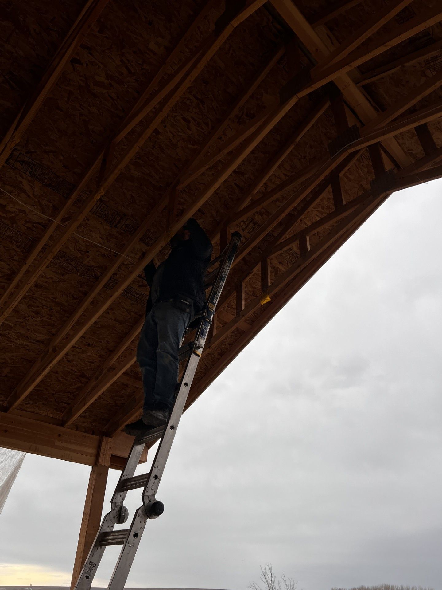 Person on a ladder, installing something on the underside of a brown wooden roof. Overcast sky in the background.