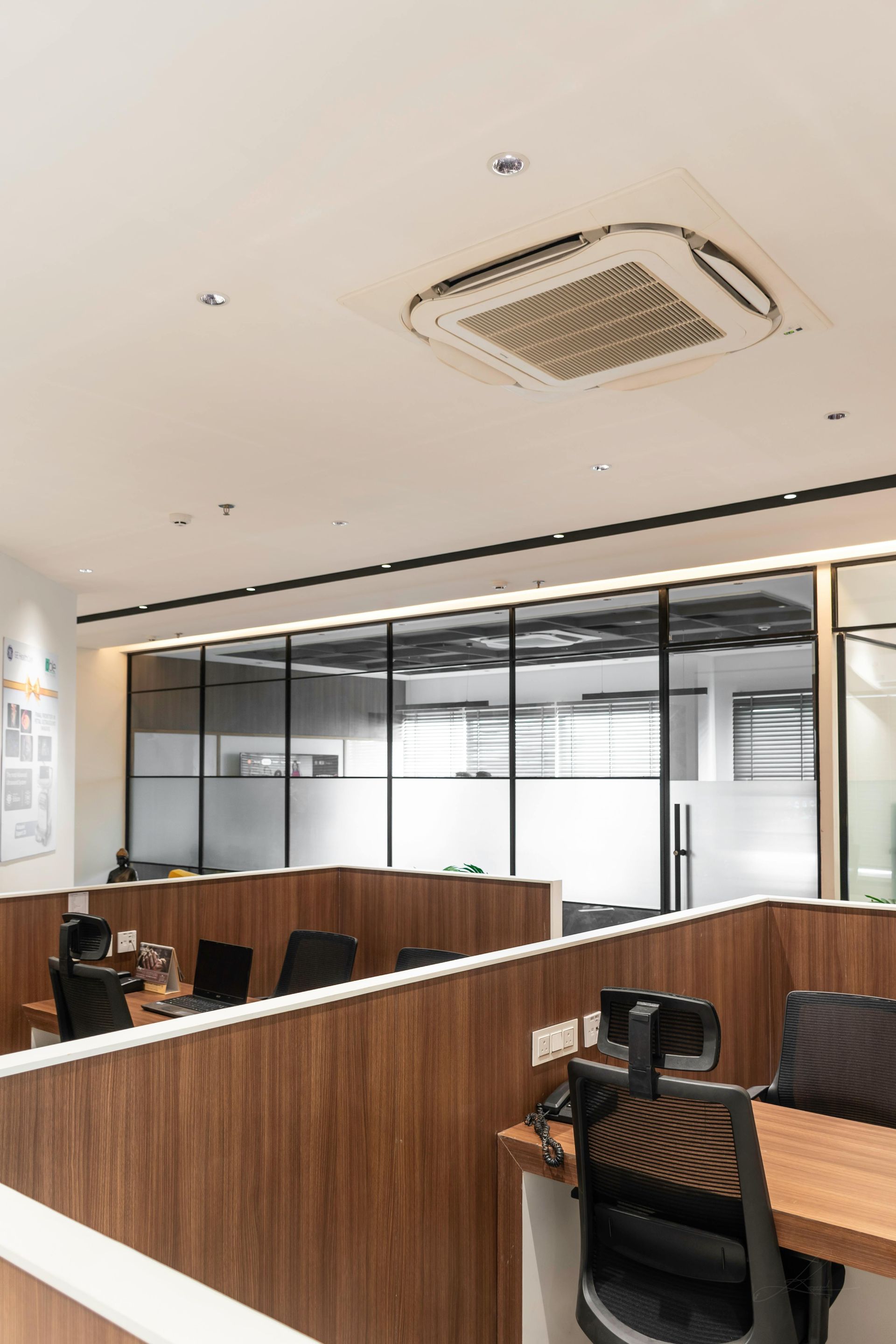Modern office interior with wood desks, black chairs, glass partitions, and a ceiling AC vent