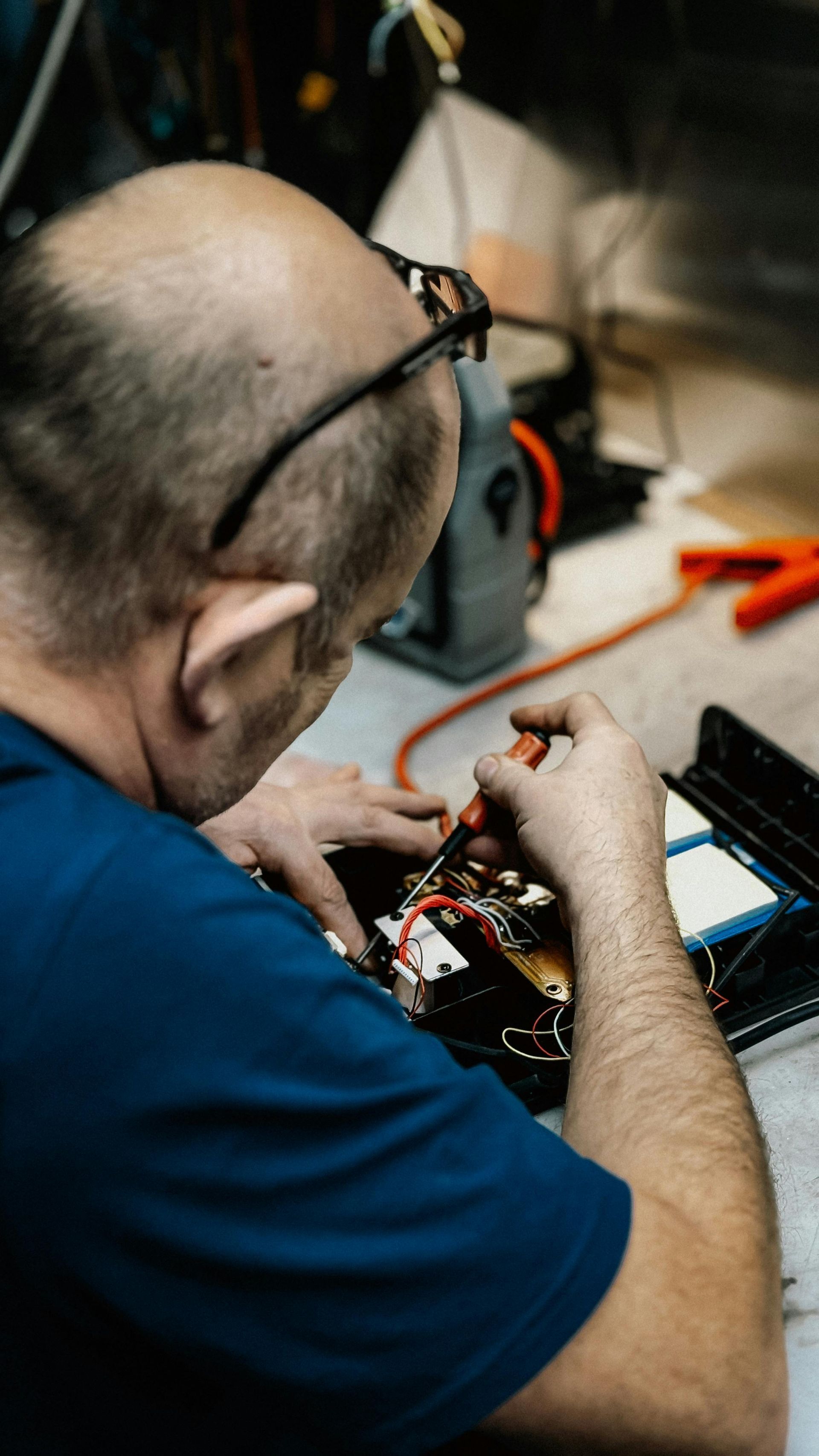 Technician repairing electronics at a workbench, viewed from above, with tools and exposed circuit board.