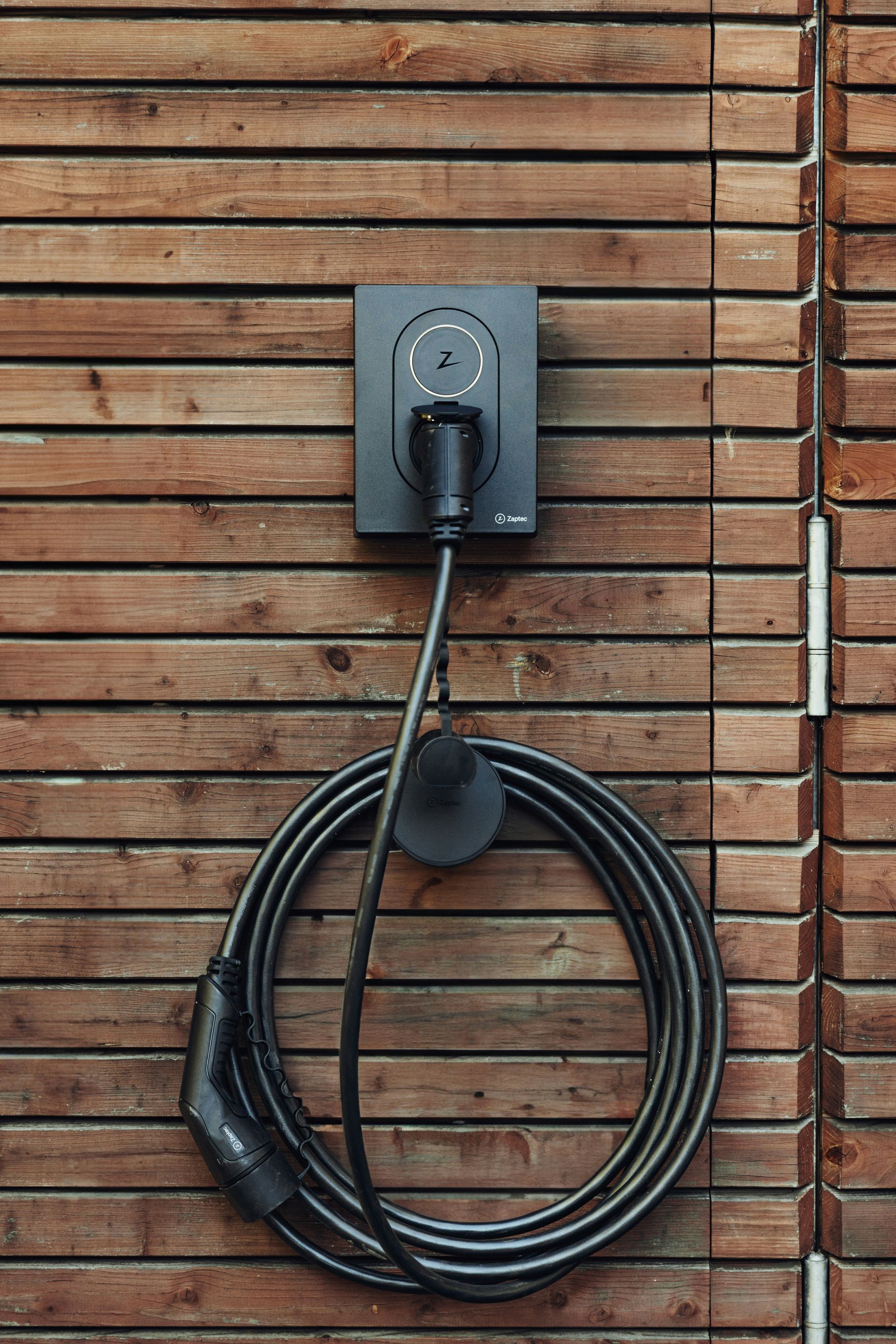 Black EV charging cable coiled beneath a wall-mounted charger on a wooden slat wall.