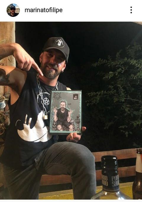 Homem de camisa preta e boné posa com uma foto emoldurada, apontando para ela ao ar livre à noite.