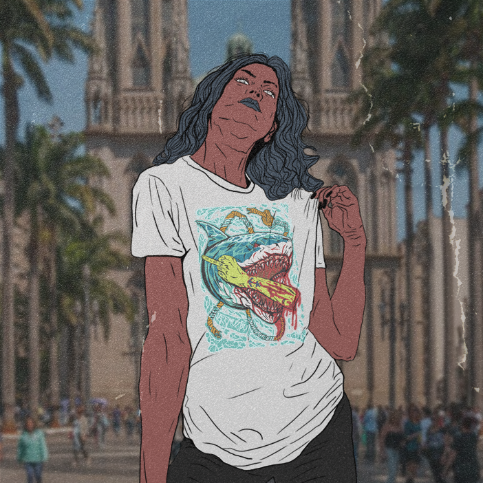 Pessoa vestindo uma camiseta branca estampada posando em frente a uma catedral com palmeiras e uma multidão.
