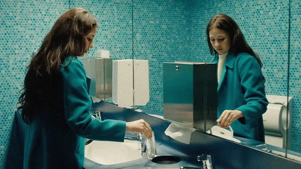 Duas mulheres com jaquetas verde-azuladas estão em um quiosque, estendendo as mãos em direção a um balcão de vidro em uma sala moderna.