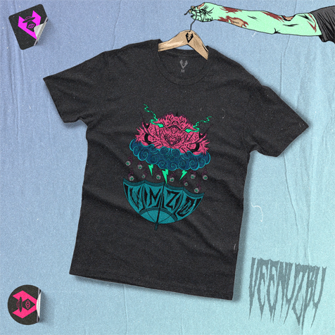 Camiseta preta com estampa floral de caveira em tons de verde-azulado e rosa, estendida sobre uma superfície azul com adesivos próximos.