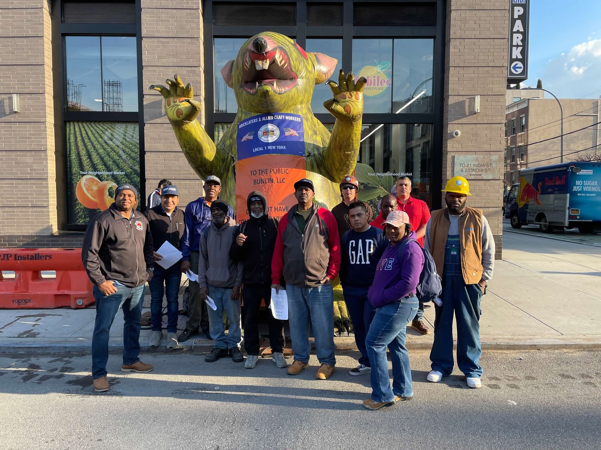 Group of people standing in front of a giant rat statue. They are wearing work clothes, and are outside a building.