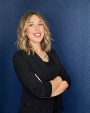 Smiling person in a black outfit with arms crossed against a blue wall.