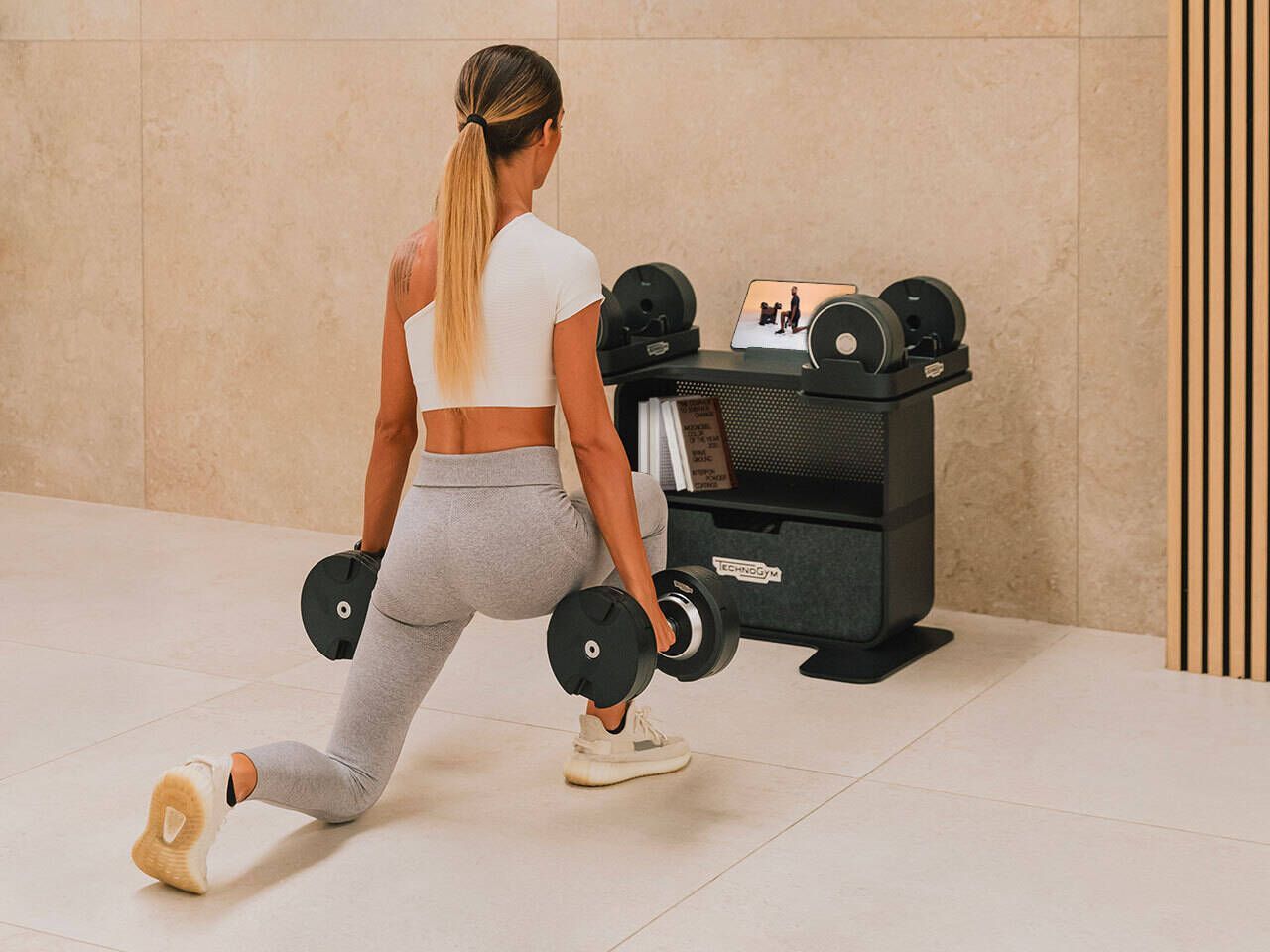 A person performing a weighted lunge in a minimalist room with a dumbbell rack.