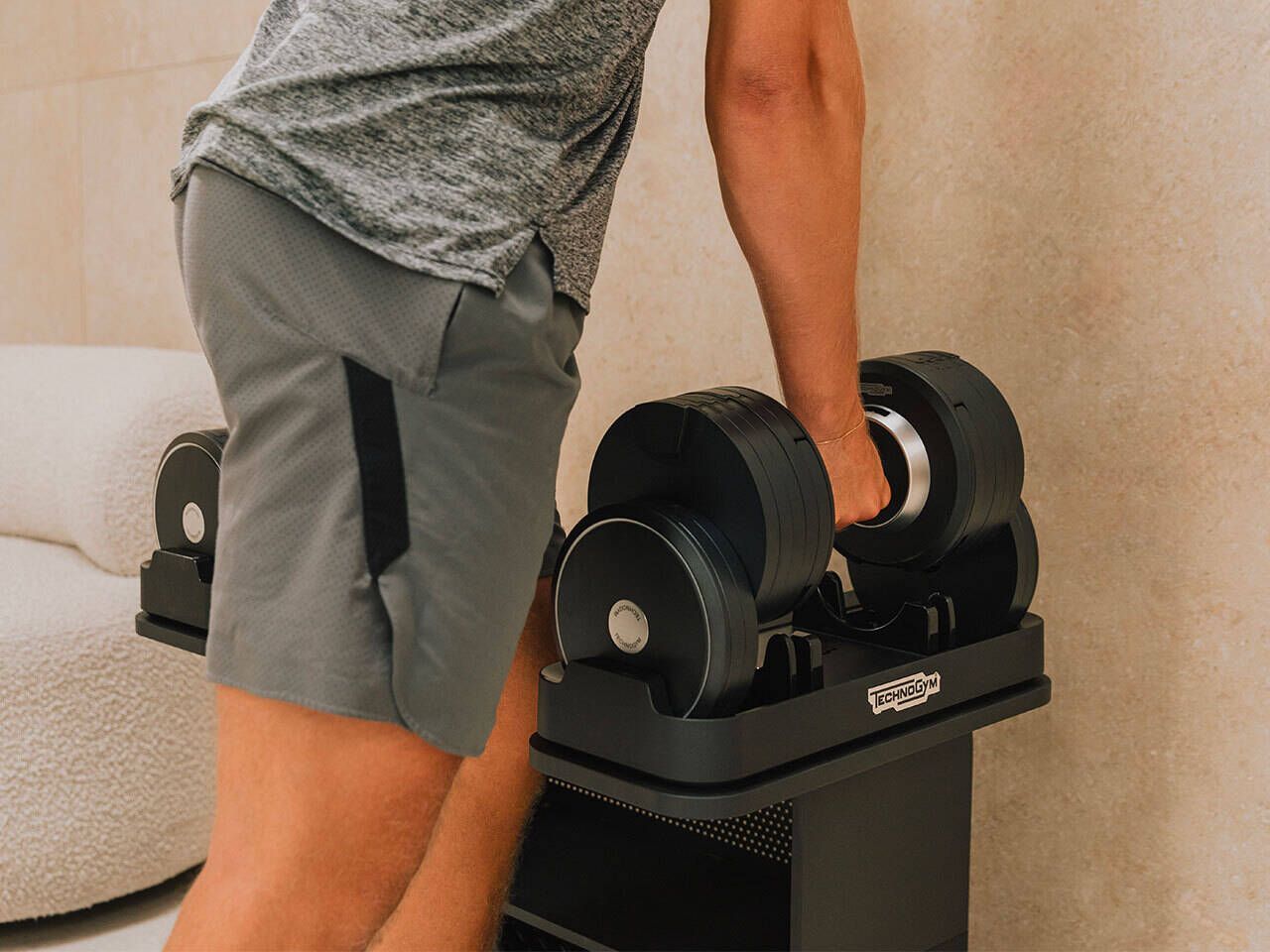 A person in workout attire lifting a black adjustable dumbbell from its storage stand in a room.