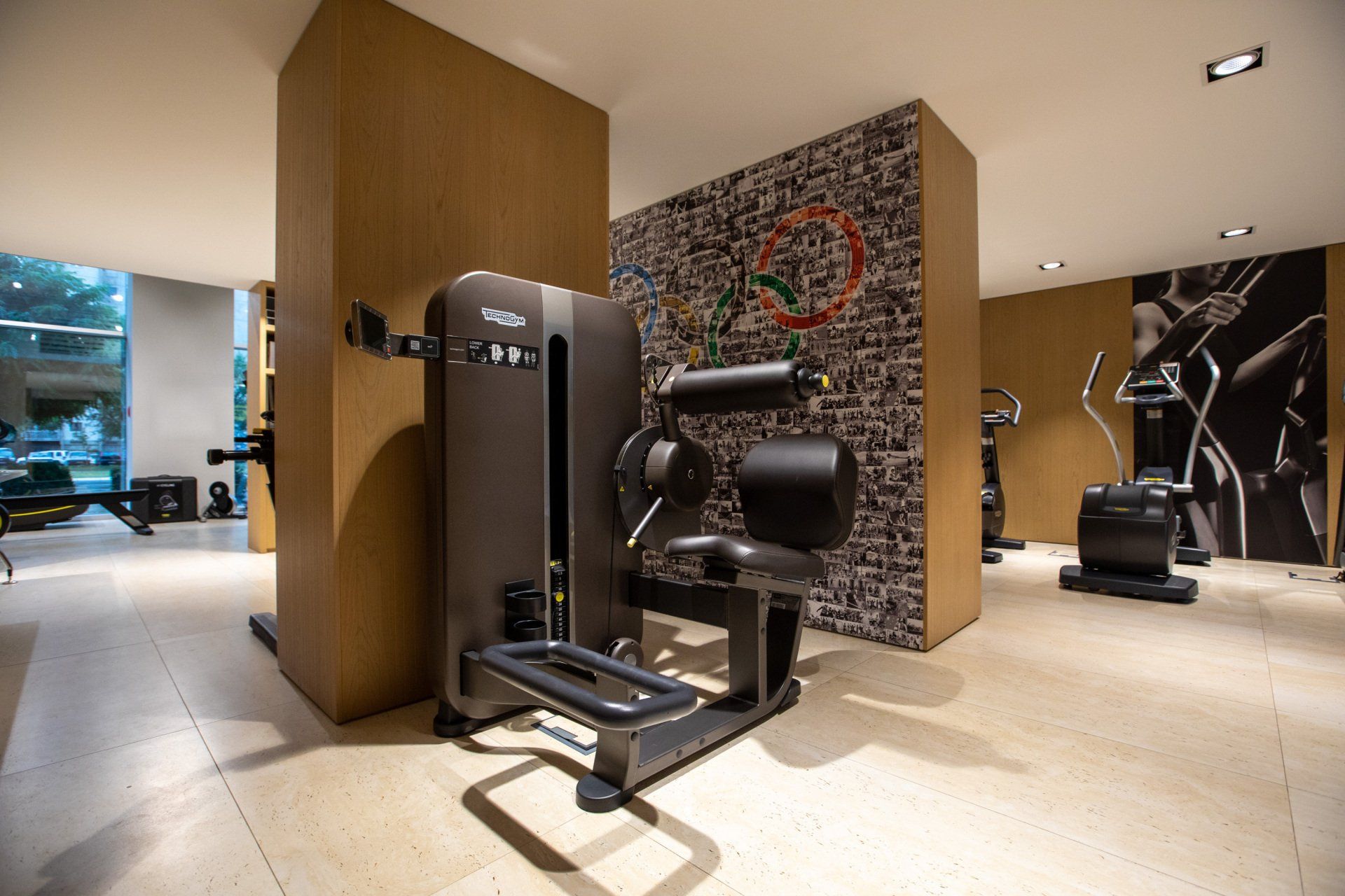 Модерна унутрашњост теретане са Technogym опремом за вежбање, стубовима обложеним дрветом и акцентним зидом са шарама.