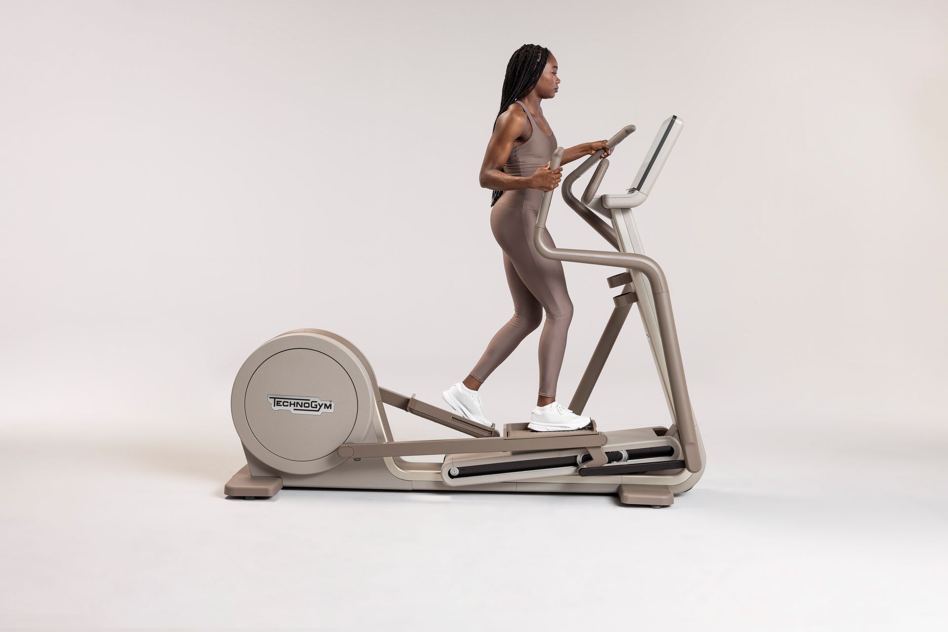 Особа вежба на беж Technogym елиптичном тренажеру на обичној, светлој позадини.