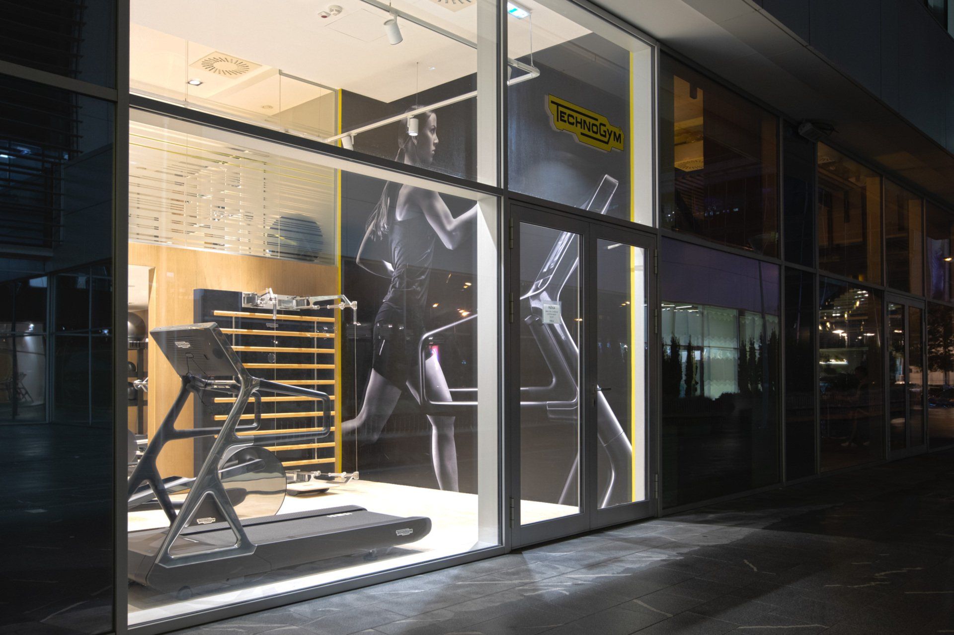 Излог продавнице Technogym ноћу, са траком за трчање и фитнес опремом видљивом кроз стакло од пода до плафона.