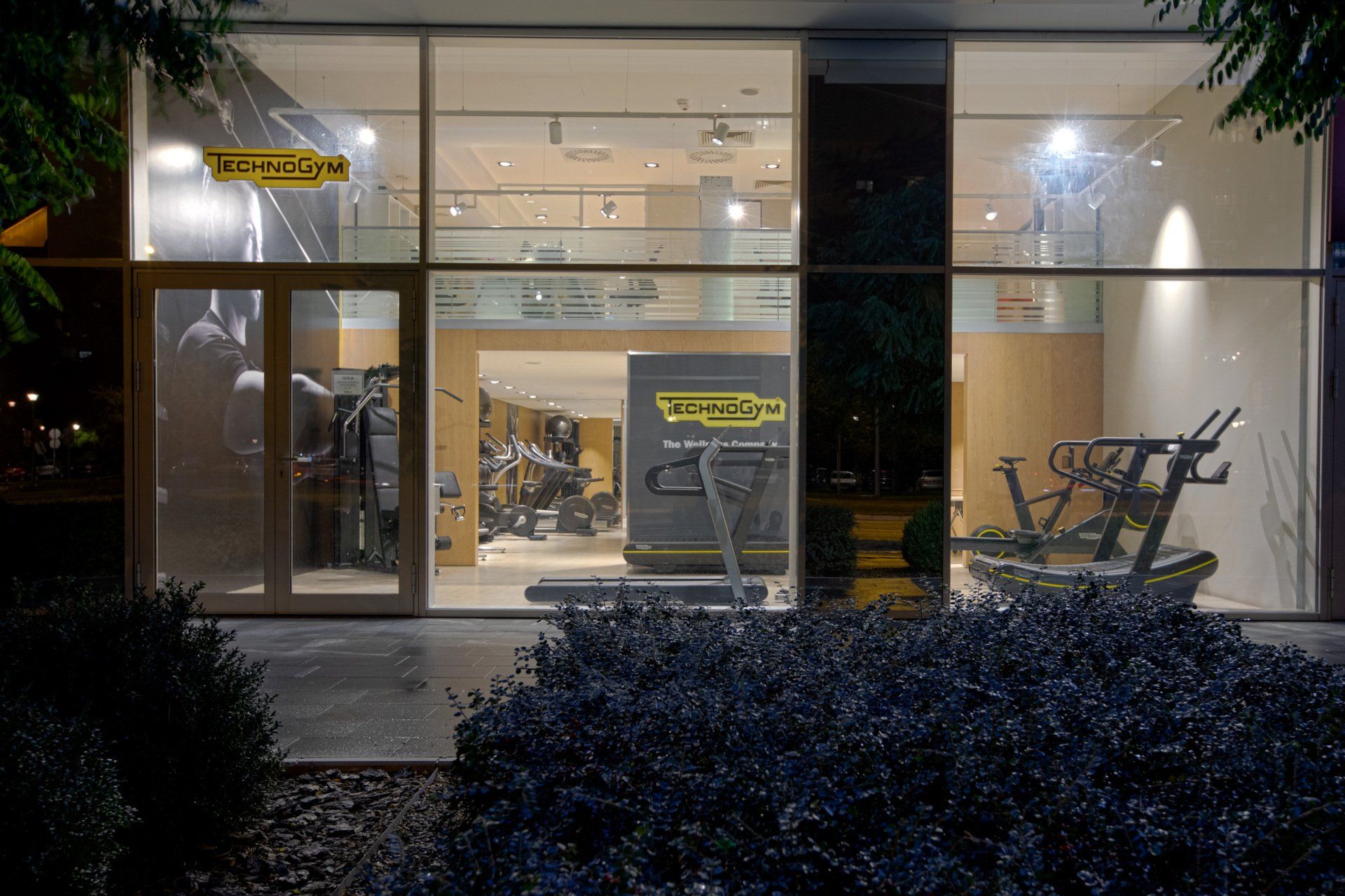 Светло осветљен салон за фитнес центар Technogym виђен кроз велики стаклени излог ноћу, са опремом за вежбање.
