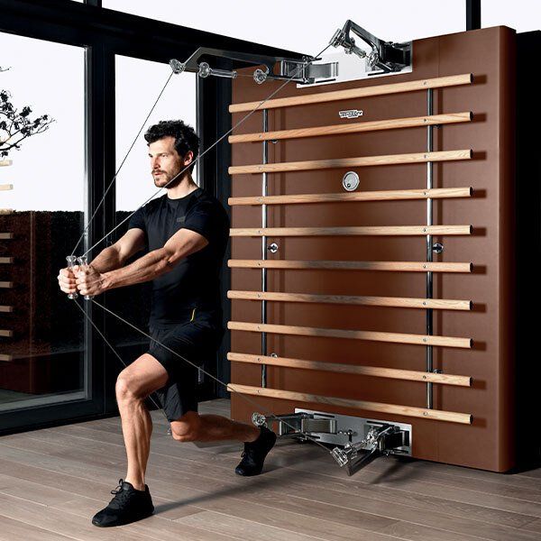 A person performing a lunge while using a wooden wall-mounted cable exercise machine in a bright, modern room.