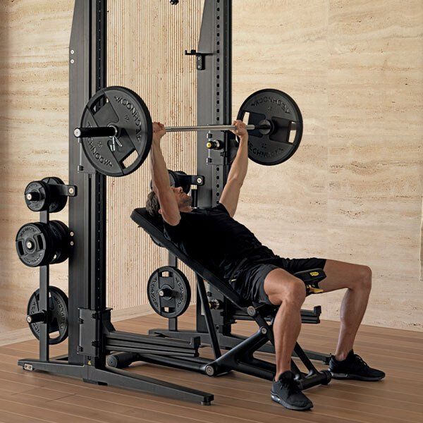 A person performs an incline bench press using a black barbell and rack in a gym with light-colored wood walls.