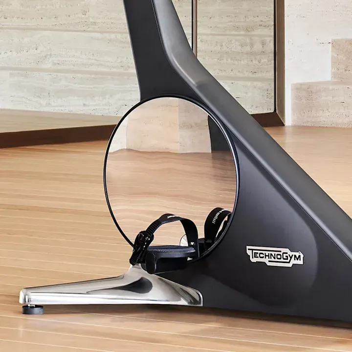 Technogym stationary bike base featuring a circular cutout, chrome foot, and black frame on a wooden floor.