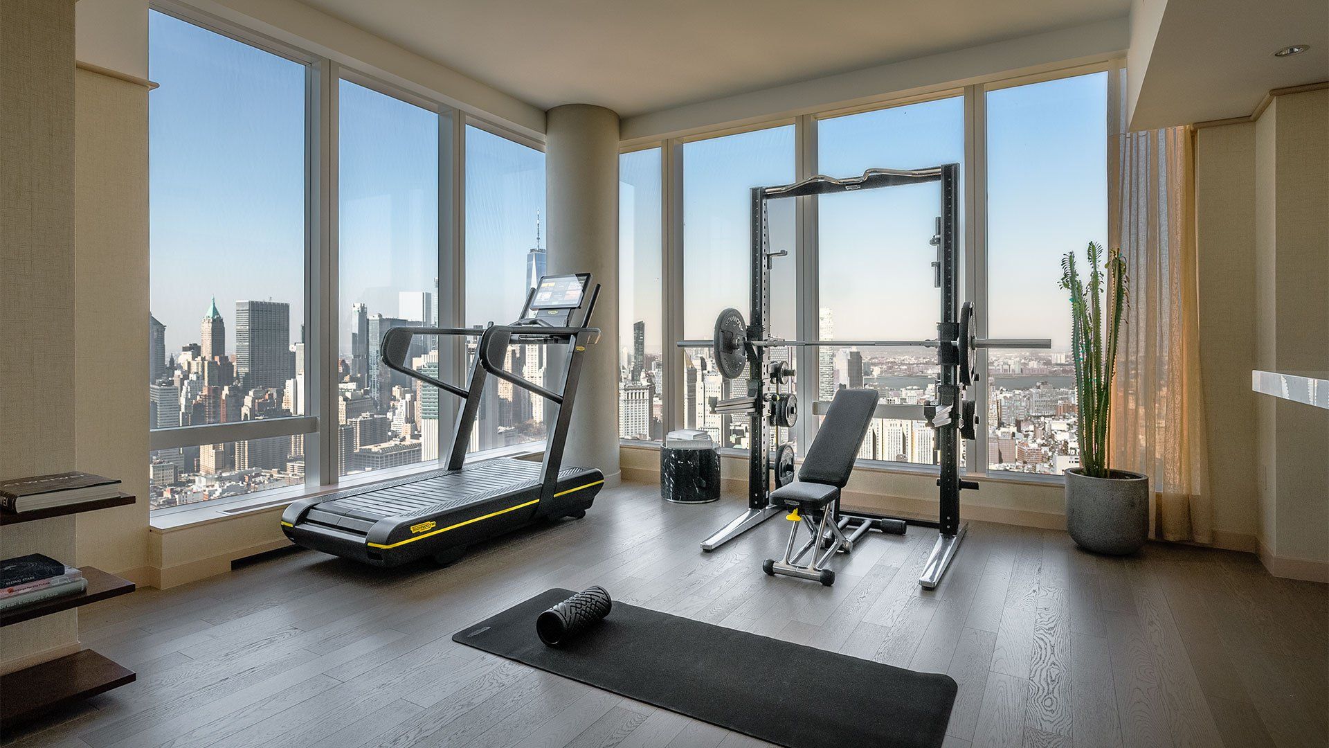 A home gym featuring a treadmill, a weight rack with a bench, and a yoga mat, positioned before floor-to-ceiling windows.