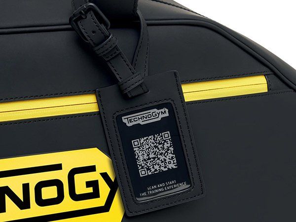 A black Technogym bag with a yellow zipper and logo, featuring a luggage tag with a QR code and instructions to scan.