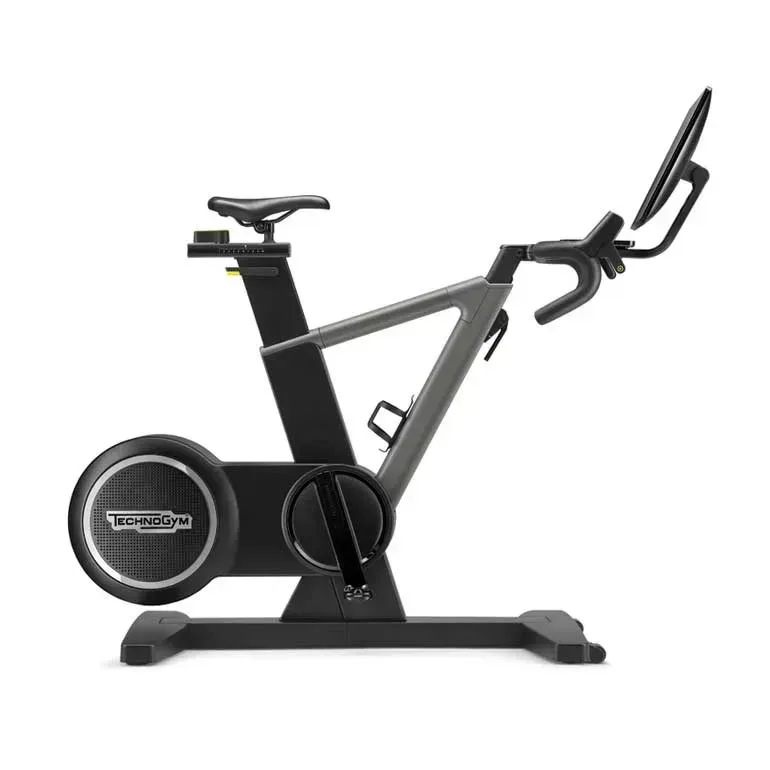 A studio shot of a black and grey Technogym stationary exercise bike with a built-in tablet display on a white background.