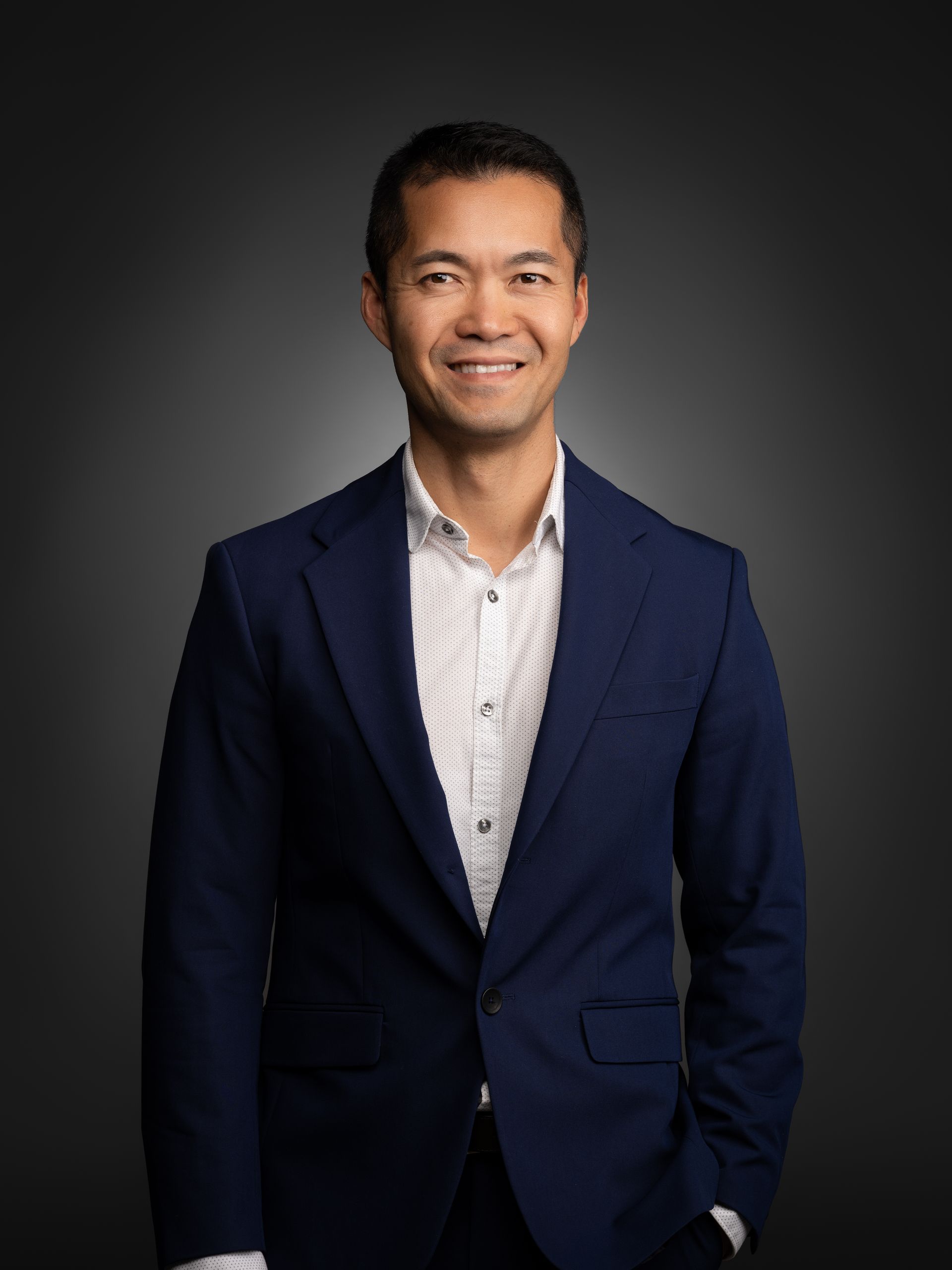Studio portrait of a smiling man in a navy blazer and white shirt against a dark background