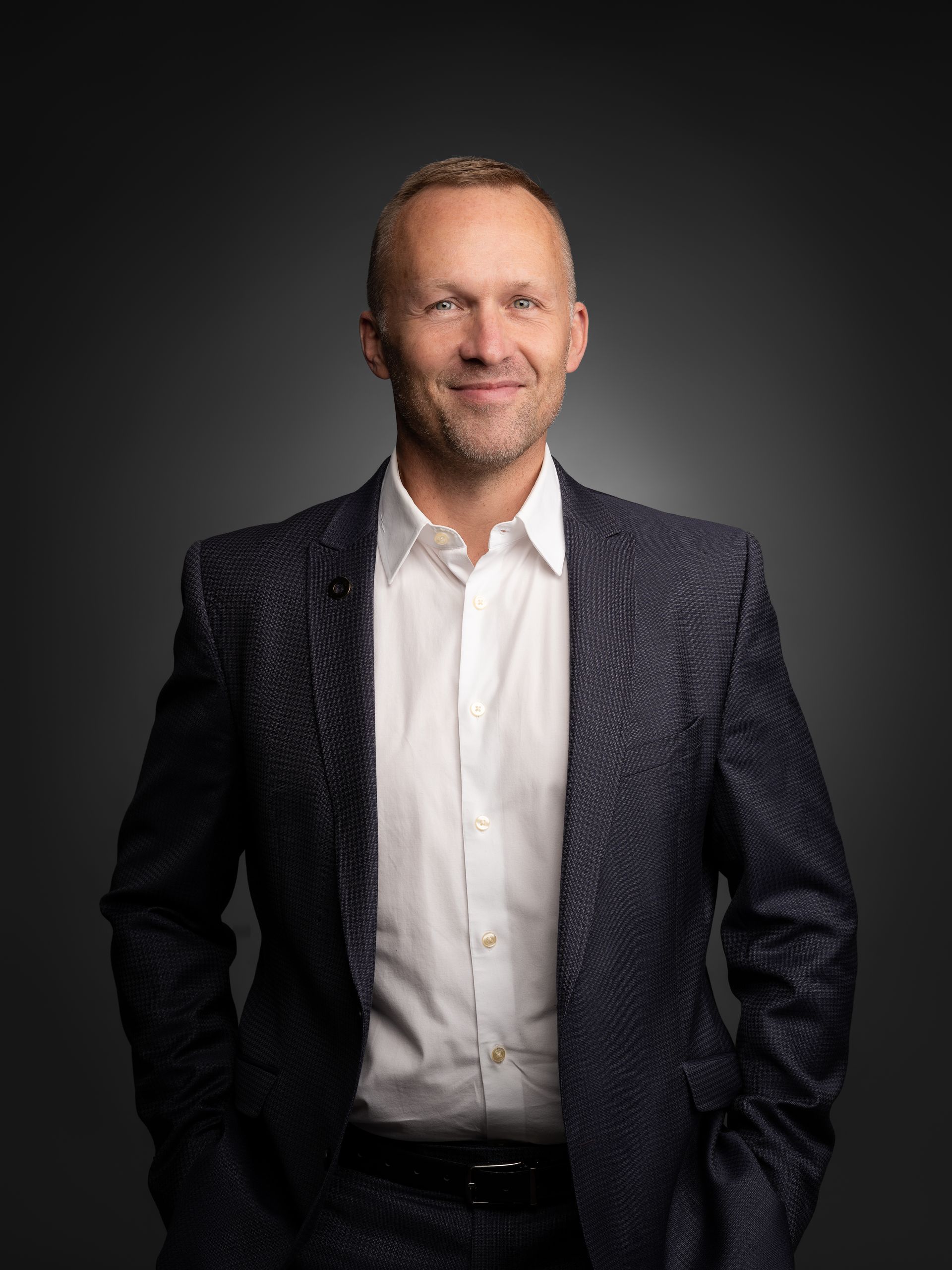 Smiling man in a dark suit and white shirt standing against a dark studio background
