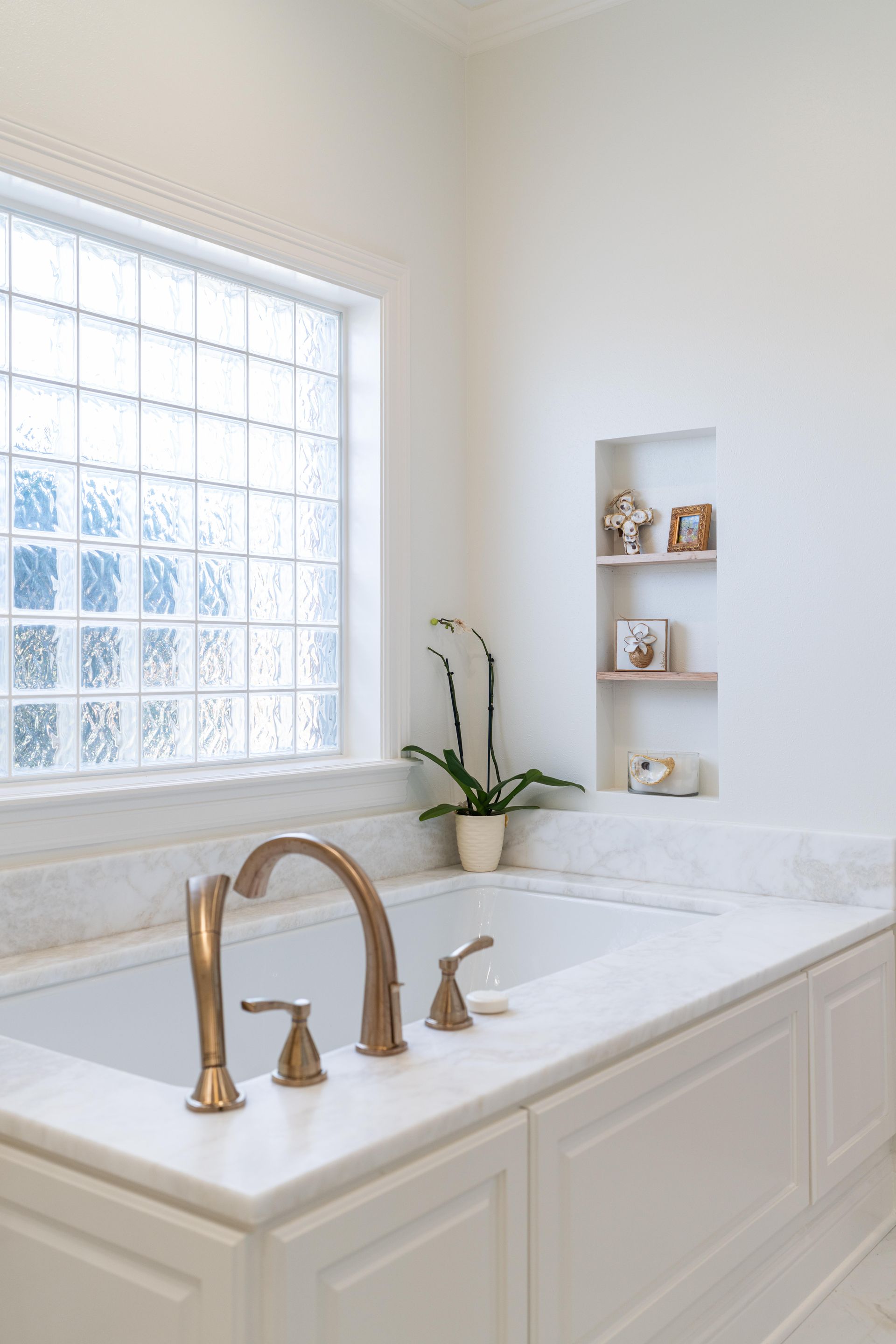 Bright bathroom vanity with brass faucet, marble countertop, and a window beside built-in wall shelves.