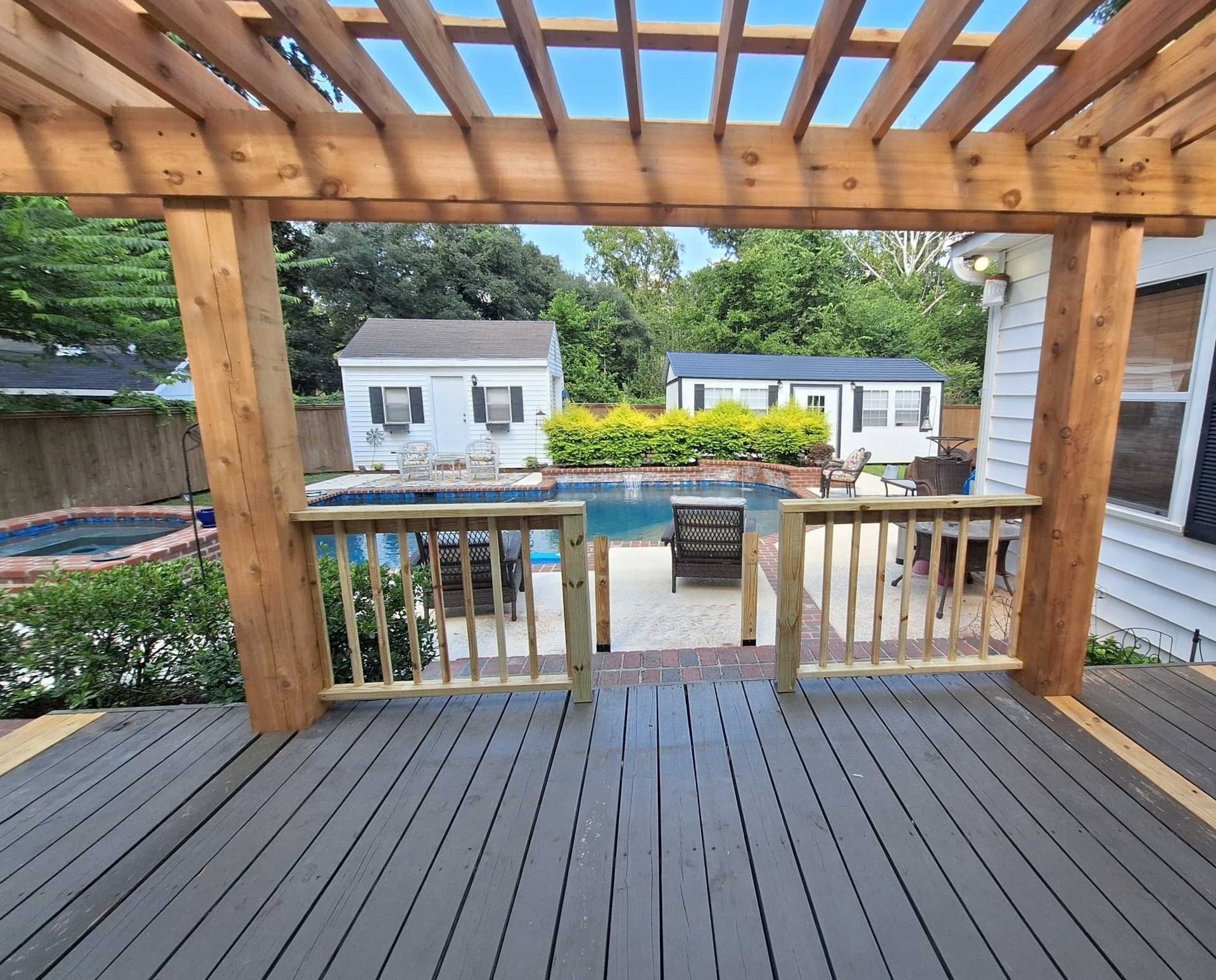 Wooden deck with pergola overlooking a backyard patio and two small buildings beyond the fence