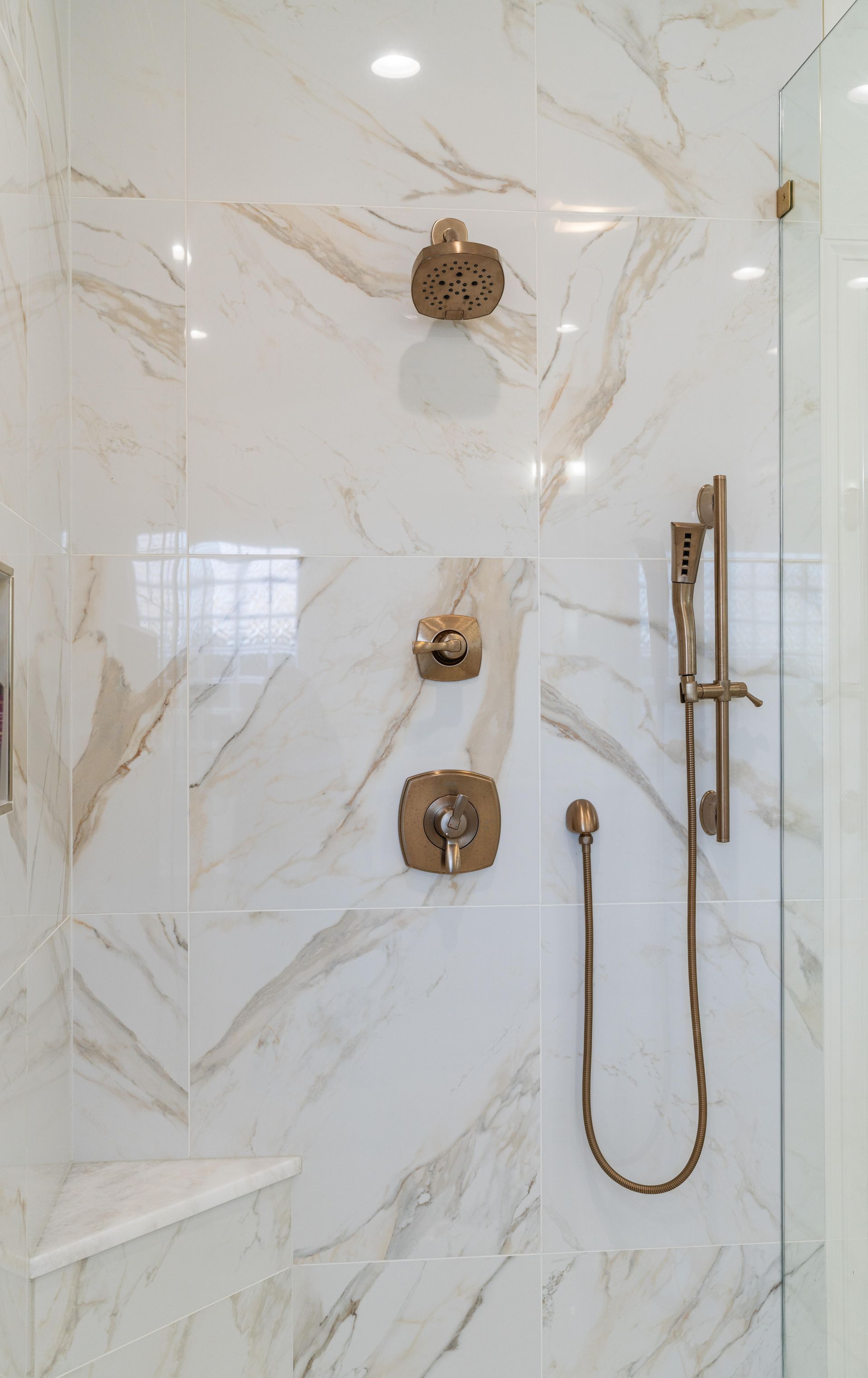 Marble-tiled shower with gold fixtures, built-in shelves, and handheld showerhead.