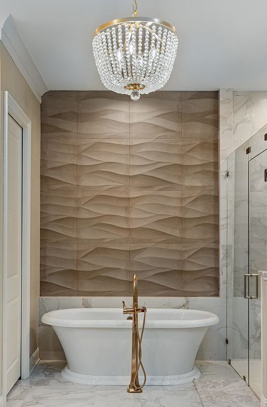 Elegant bathroom with chandelier, freestanding tub, and gold floor faucet in front of a textured tile wall