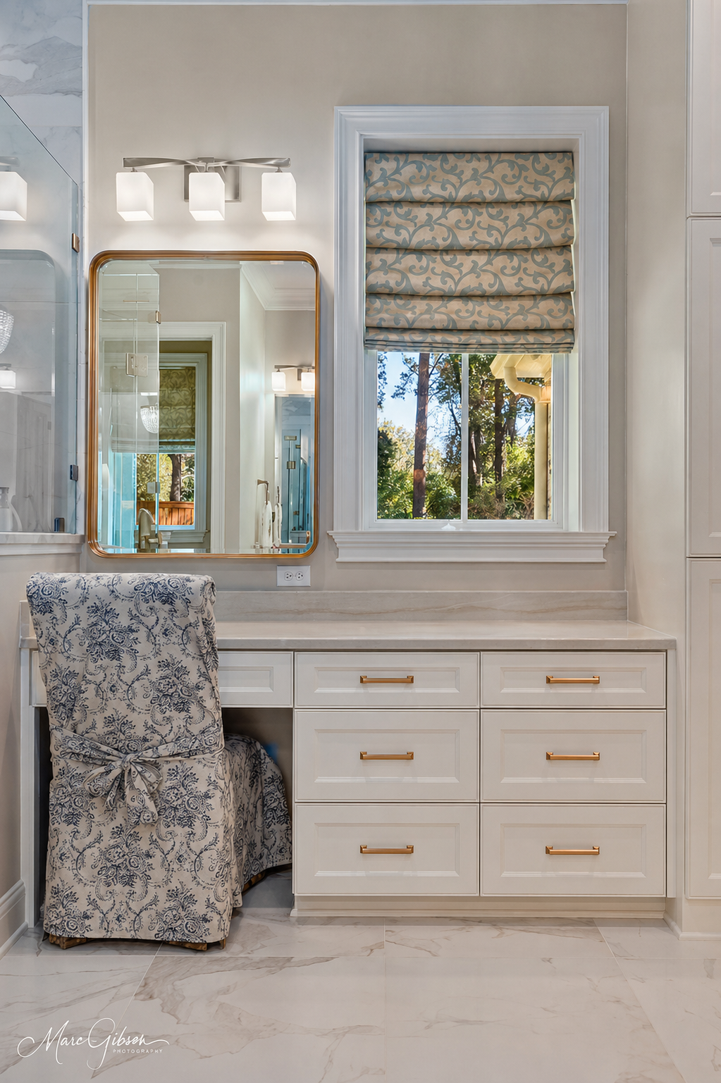 Elegant white vanity with mirror, gold hardware, patterned stool, and window with patterned Roman shade