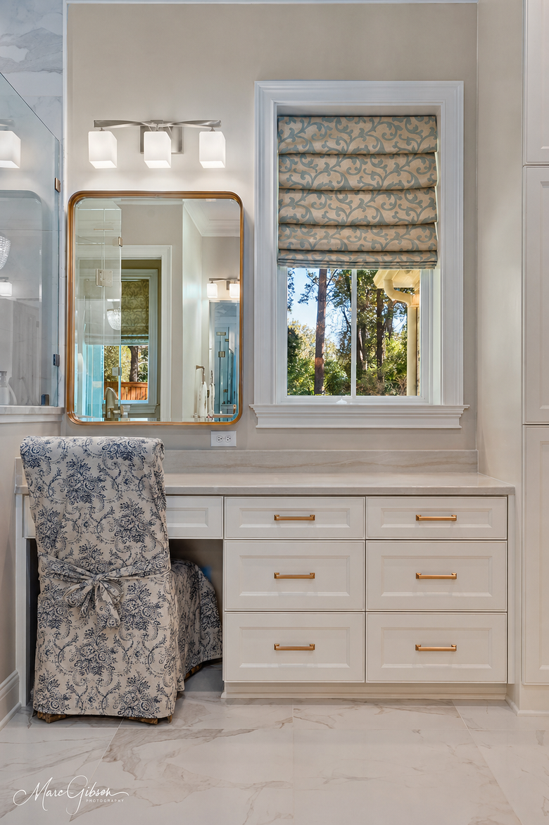 Elegant white vanity with mirror, gold hardware, patterned stool, and window with patterned Roman shade