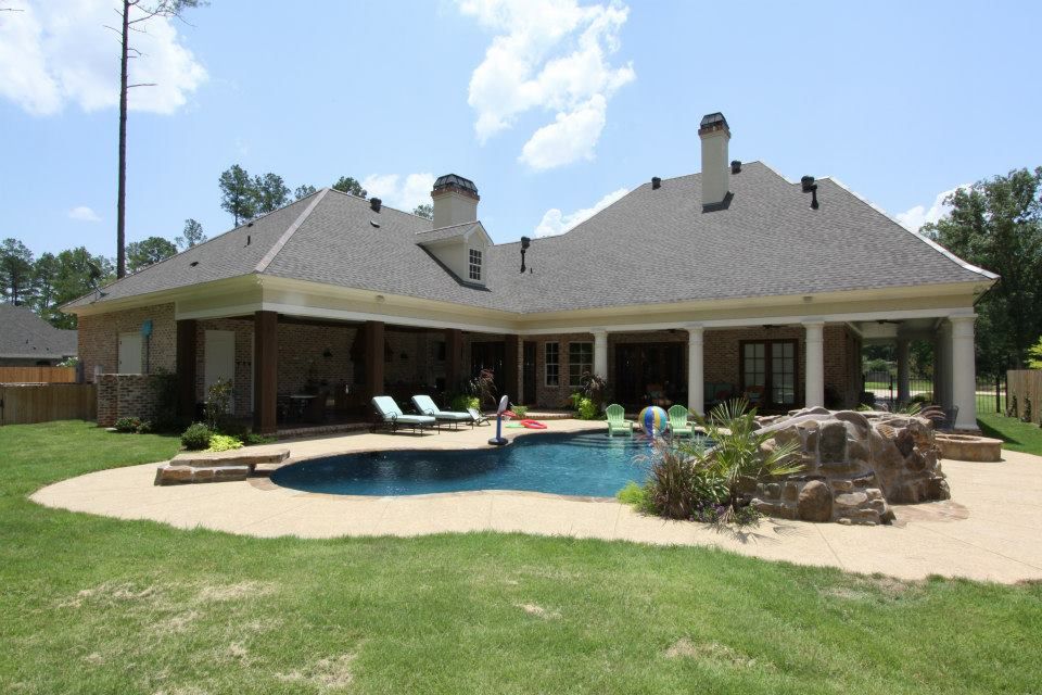 Backyard patio with a swimming pool, lounge chairs, and a large house under a bright sky