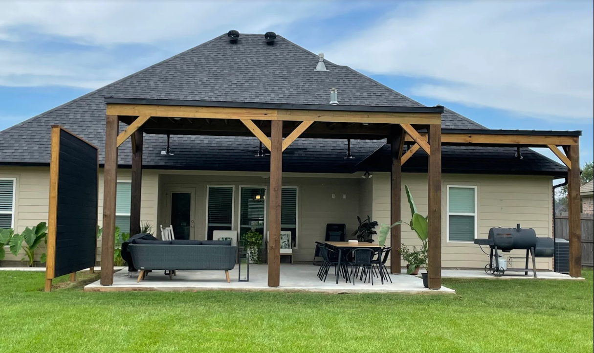 Covered patio with outdoor seating and a grill beneath a wooden pergola in a backyard