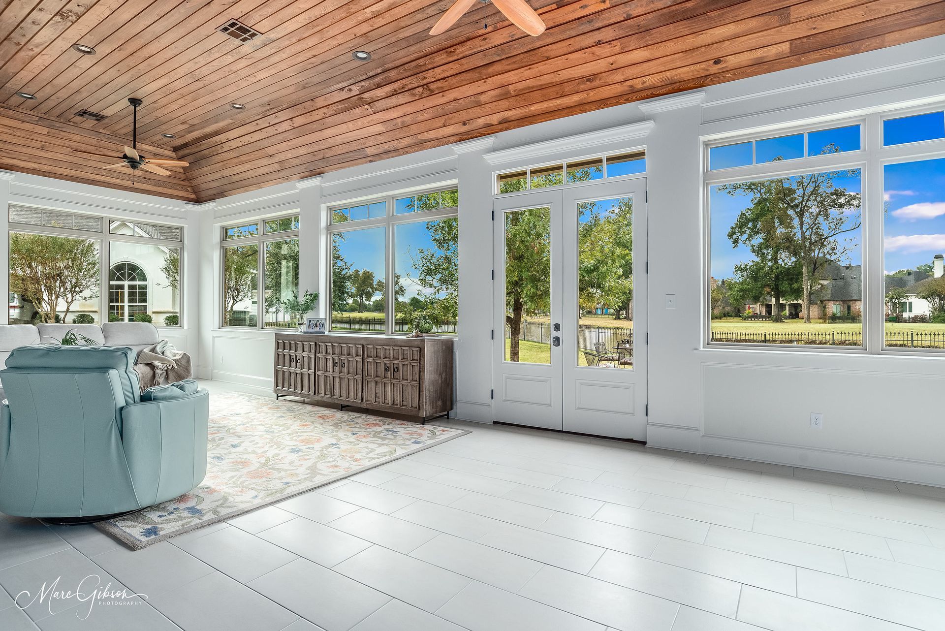 Bright sunroom with white floor, wood ceiling, large windows, and a pale blue chair by the view.
