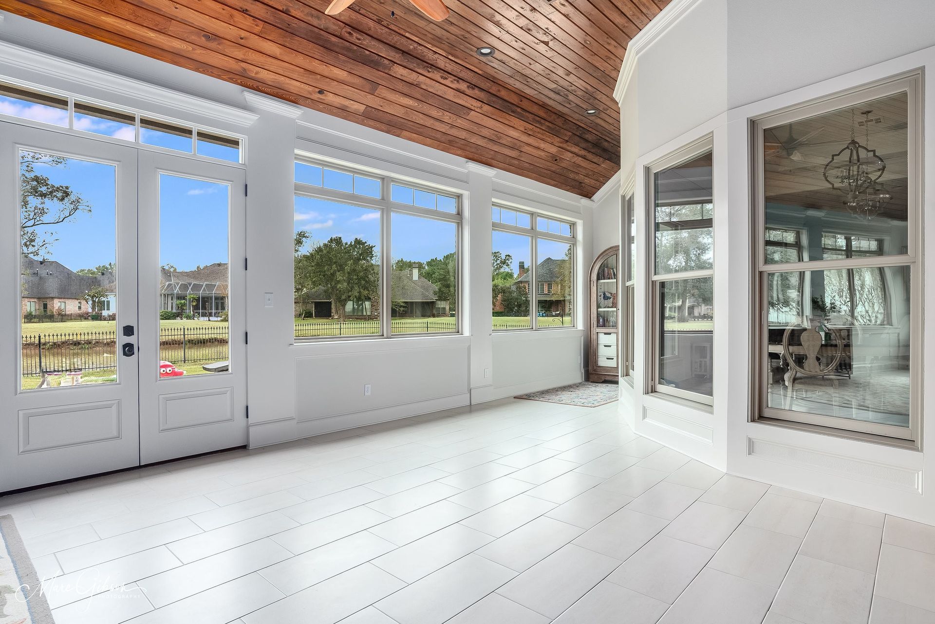 Bright sunroom with white floors, large windows, and a wood-paneled ceiling overlooking green outdoor views