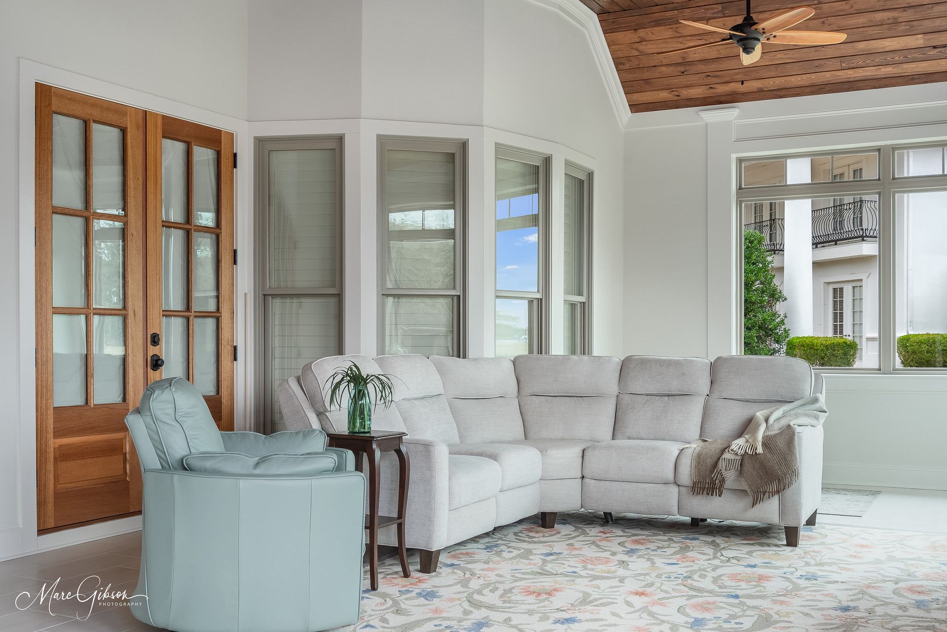 Bright white living room with a gray sectional sofa, patterned rug, wood doors, and large windows