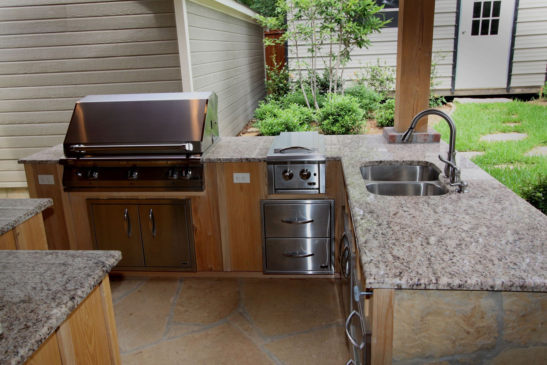 Outdoor kitchen with built-in grill, sink, and stone countertop under a covered patio.
