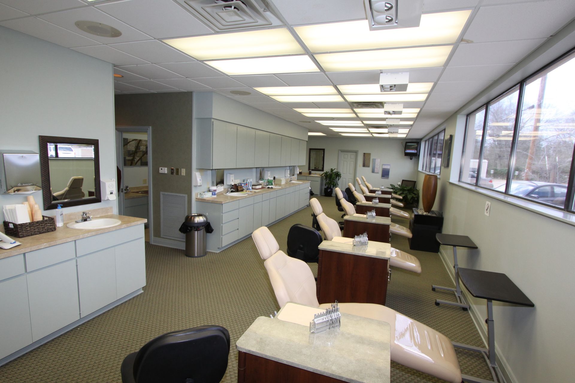 Waiting area with beige chairs, tables, and large windows along a bright office hallway.