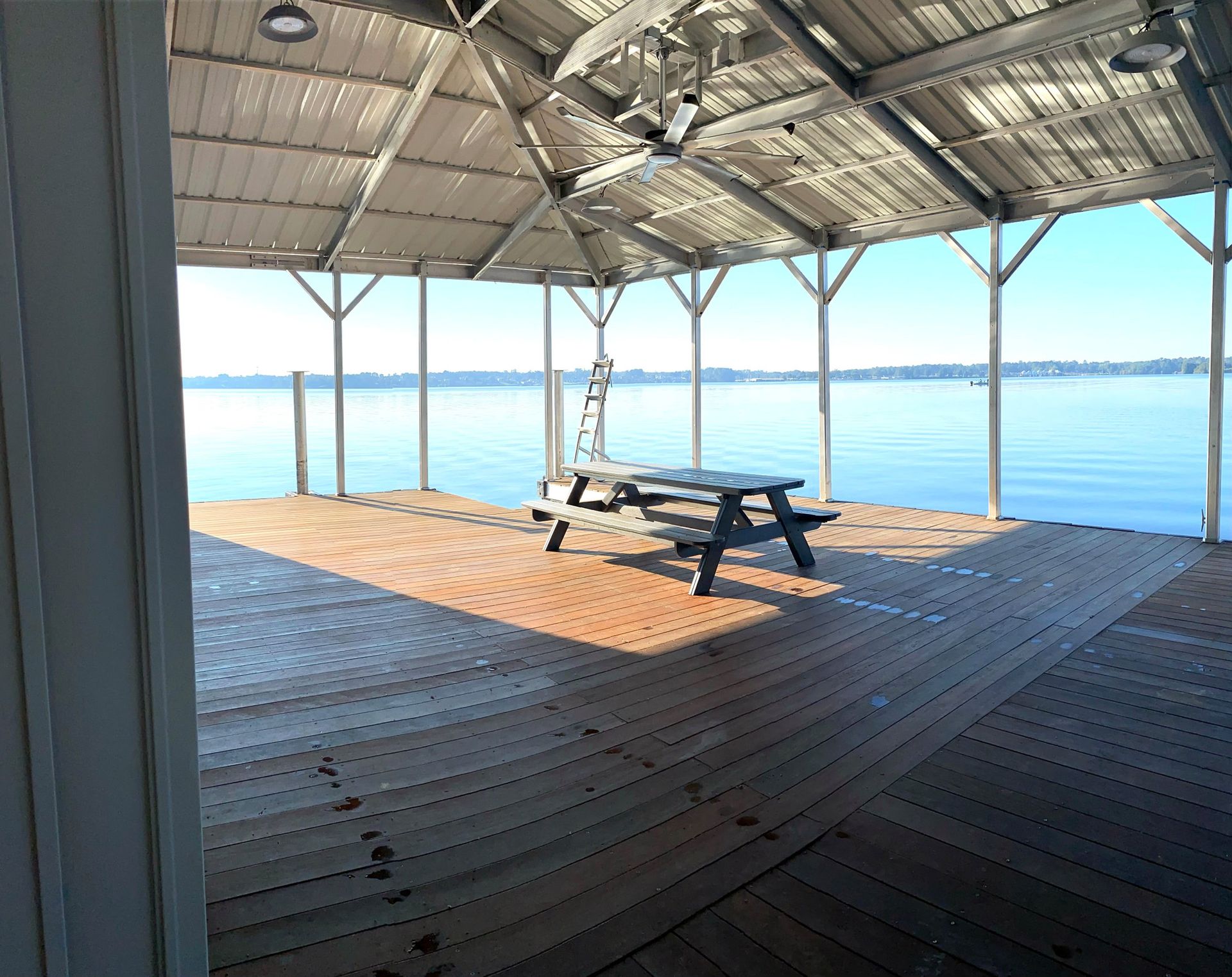 Covered wooden deck by calm water with picnic tables and bright sunlight