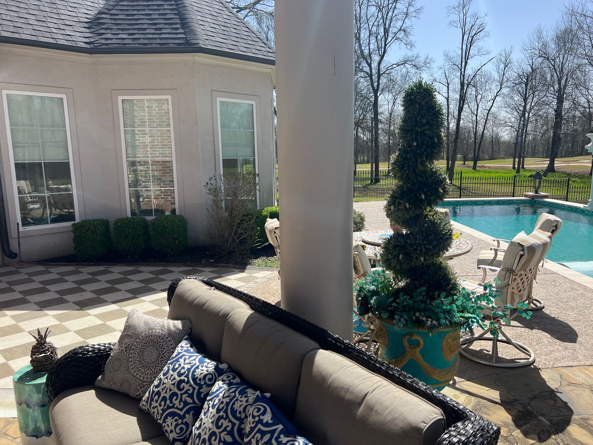 Patio with gray couch, blue patterned pillows, white column, and poolside chairs in a sunny yard