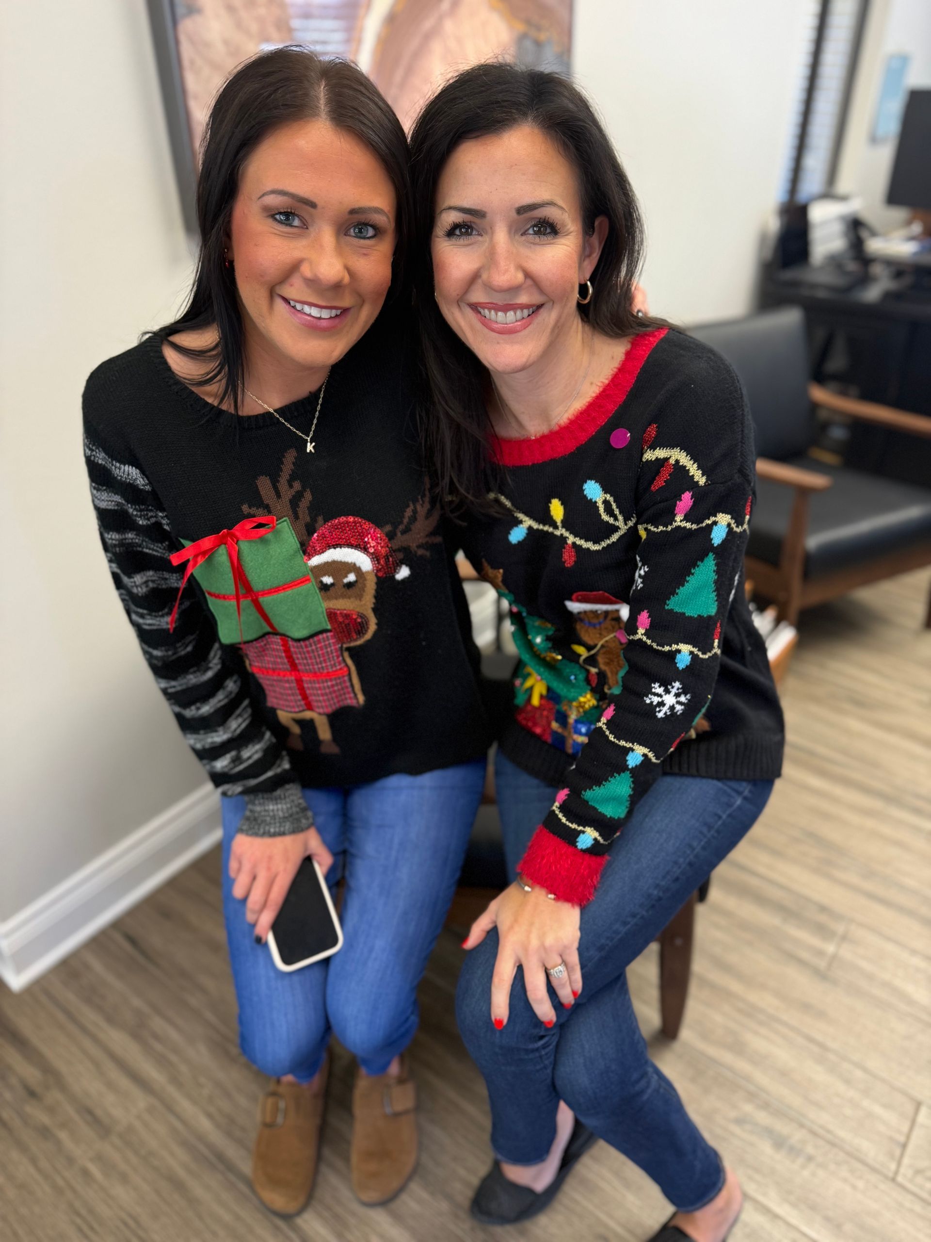 Two women smiling indoors, sitting together in festive holiday sweaters and jeans