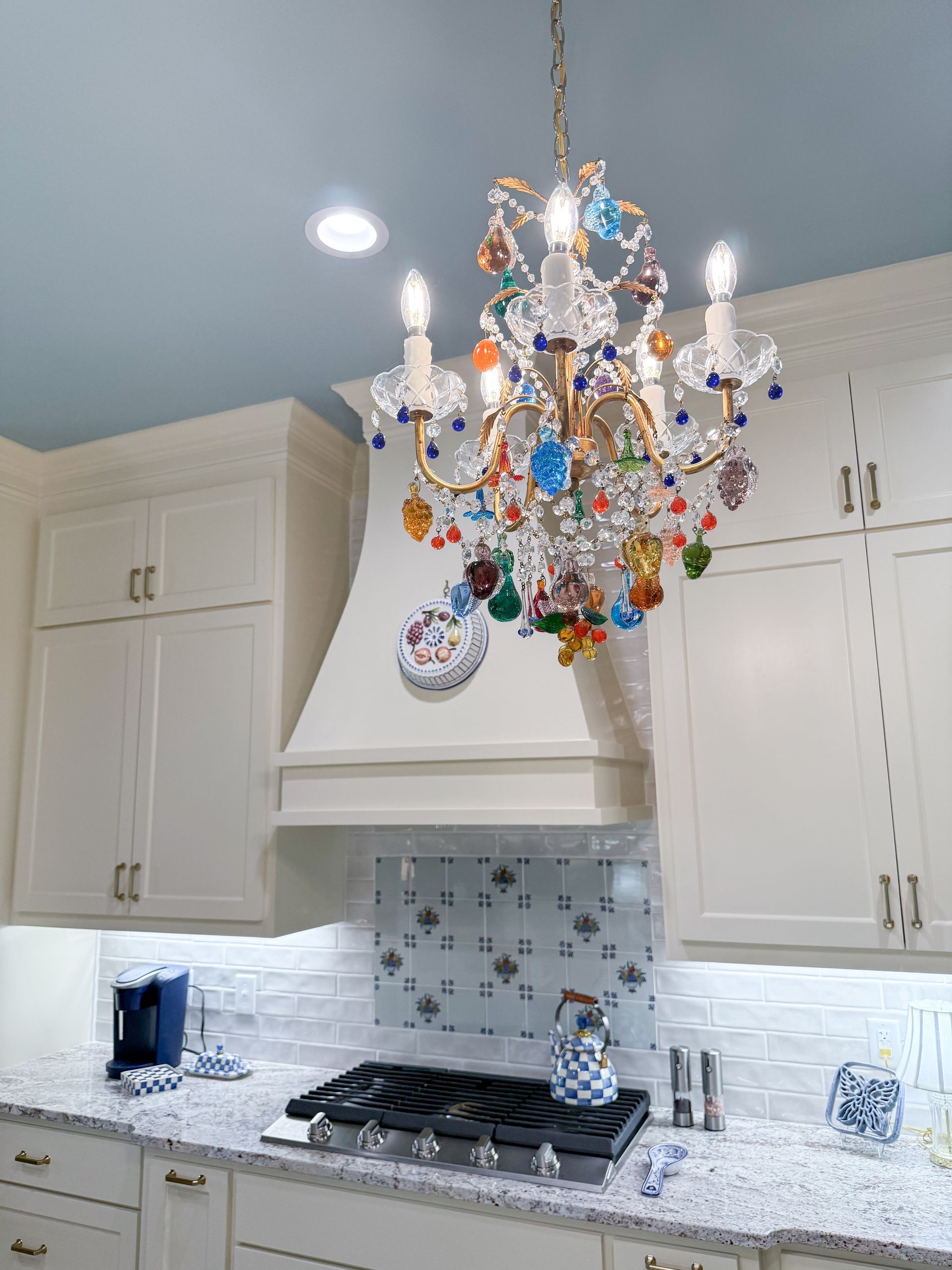 Colorful crystal chandelier hangs over a white kitchen with a gas range and mosaic tile backsplash.
