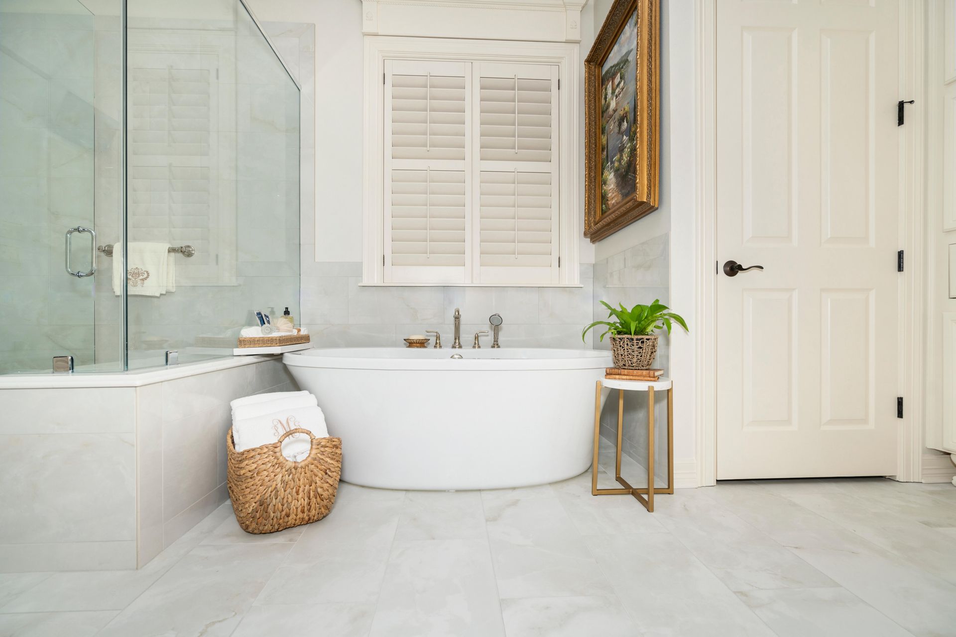Bright white bathroom with freestanding tub, glass shower, wicker basket, and plant on a small gold stand