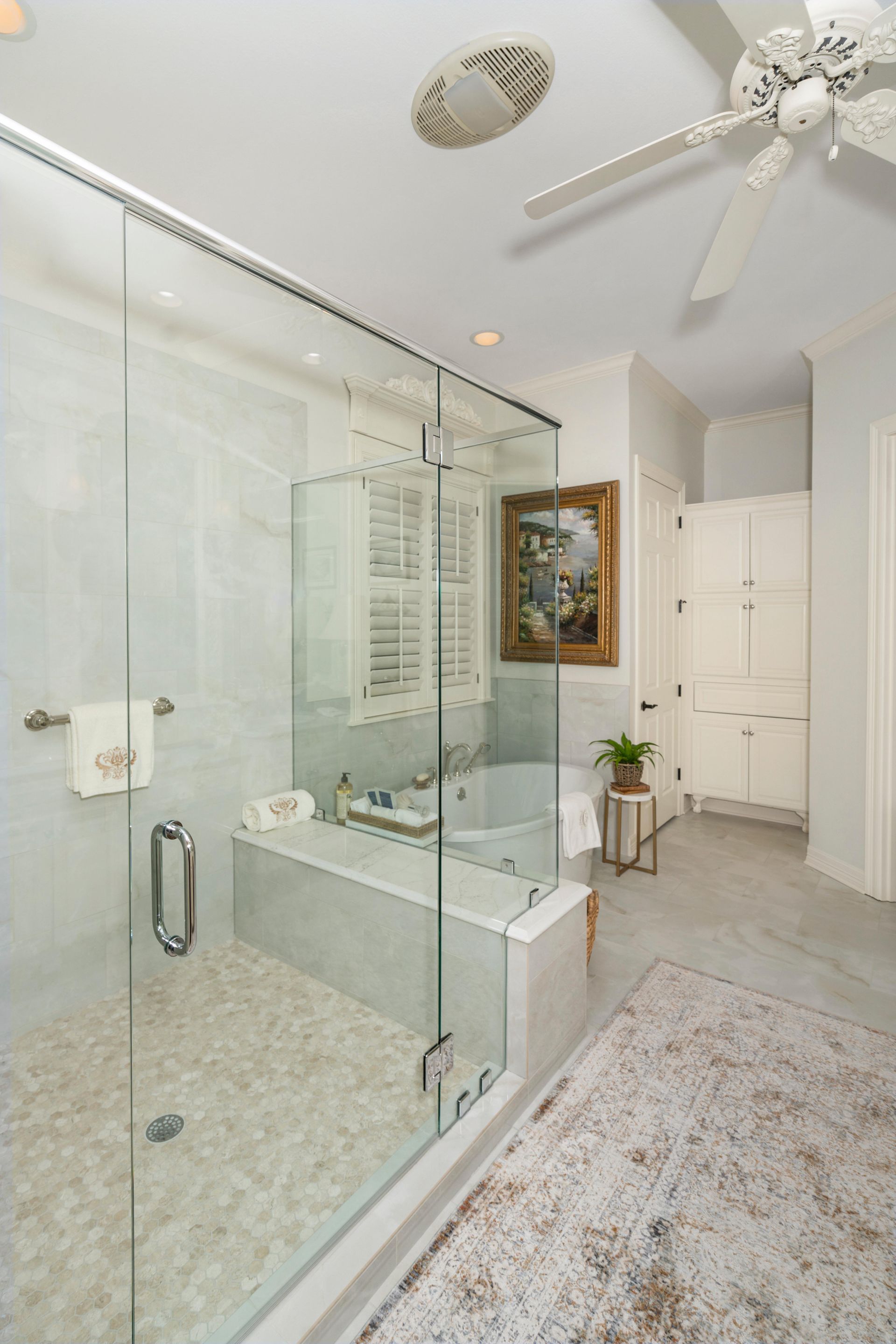 Bright modern bathroom with glass shower, freestanding tub, white cabinets, and ceiling fan