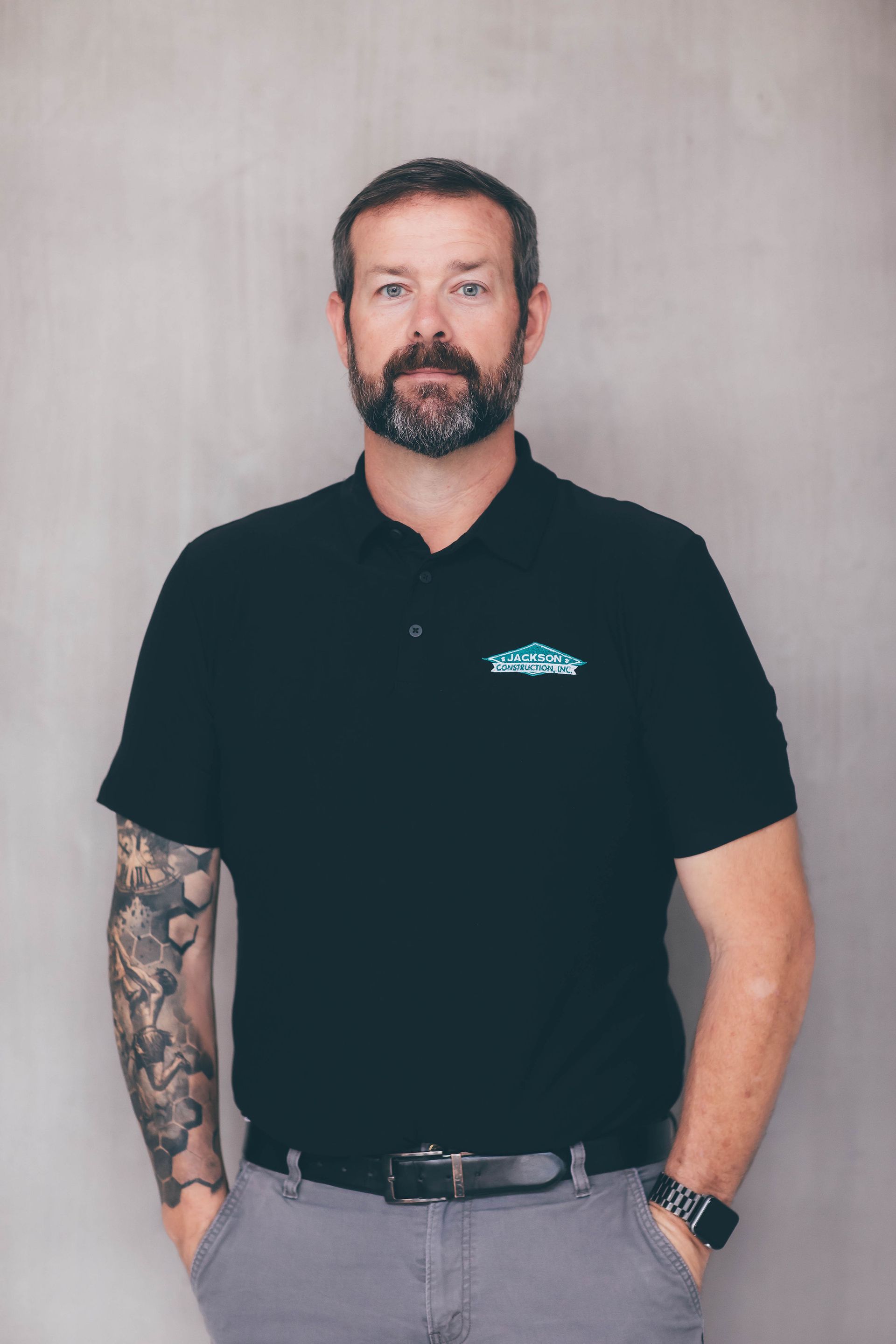 Man standing against a plain wall, wearing a black polo shirt and gray pants, with tattooed left arm and hands in pockets.