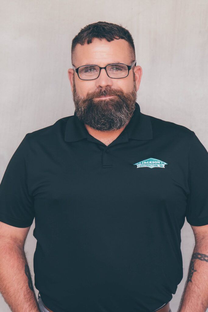 Man standing against a plain wall, wearing glasses and a black polo shirt with a logo.