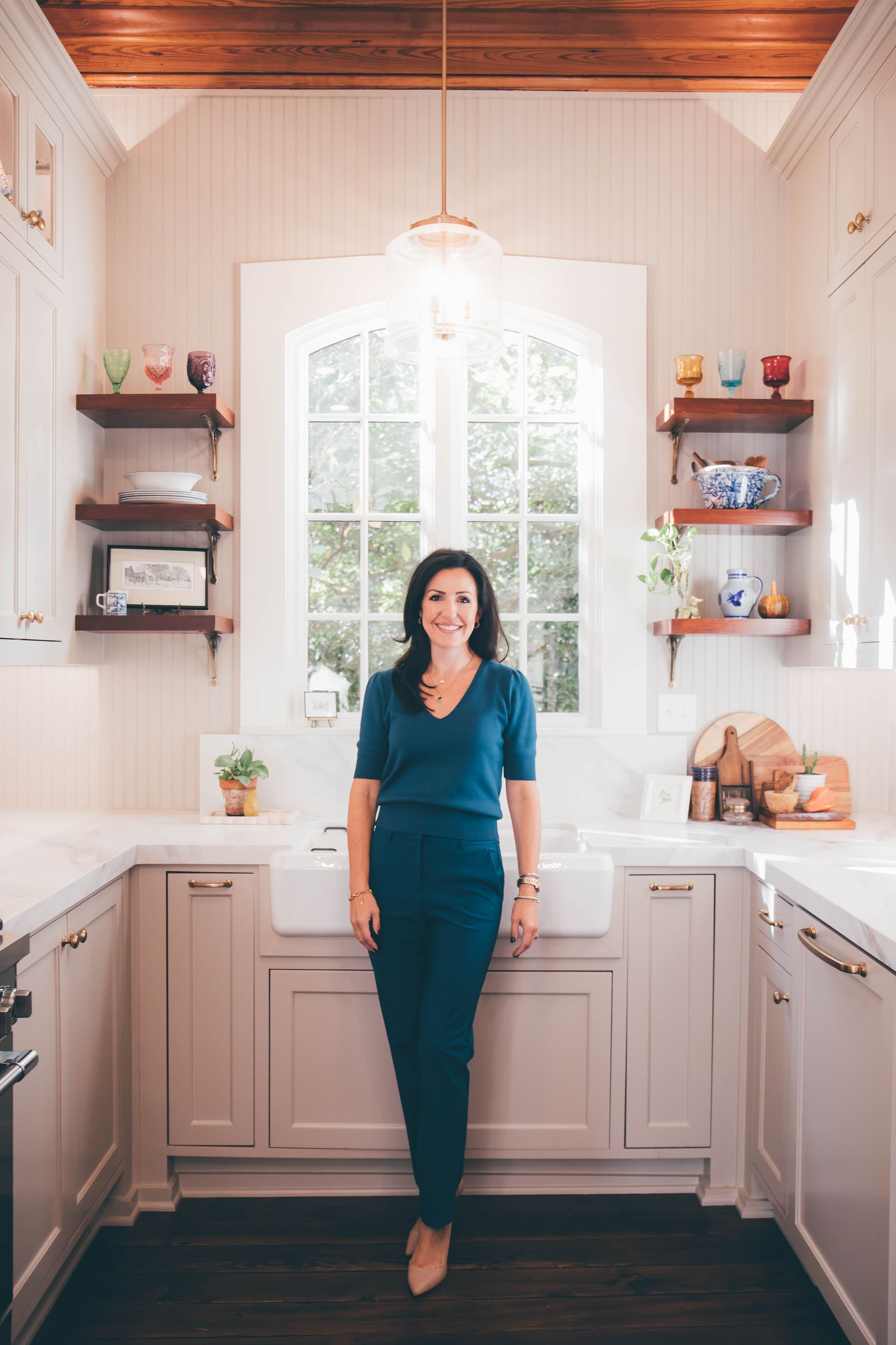 Woman standing in a bright kitchen with white cabinets and open shelving