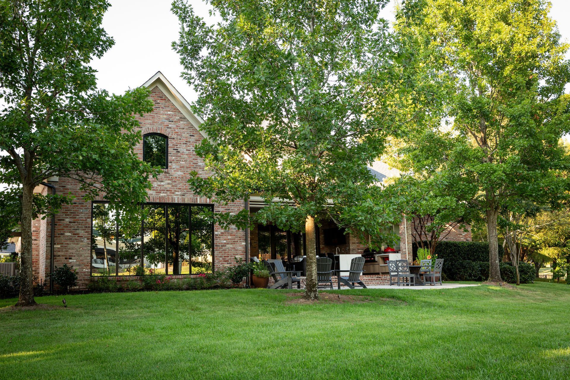 Brick building with large windows and patio seating beside a green lawn and trees
