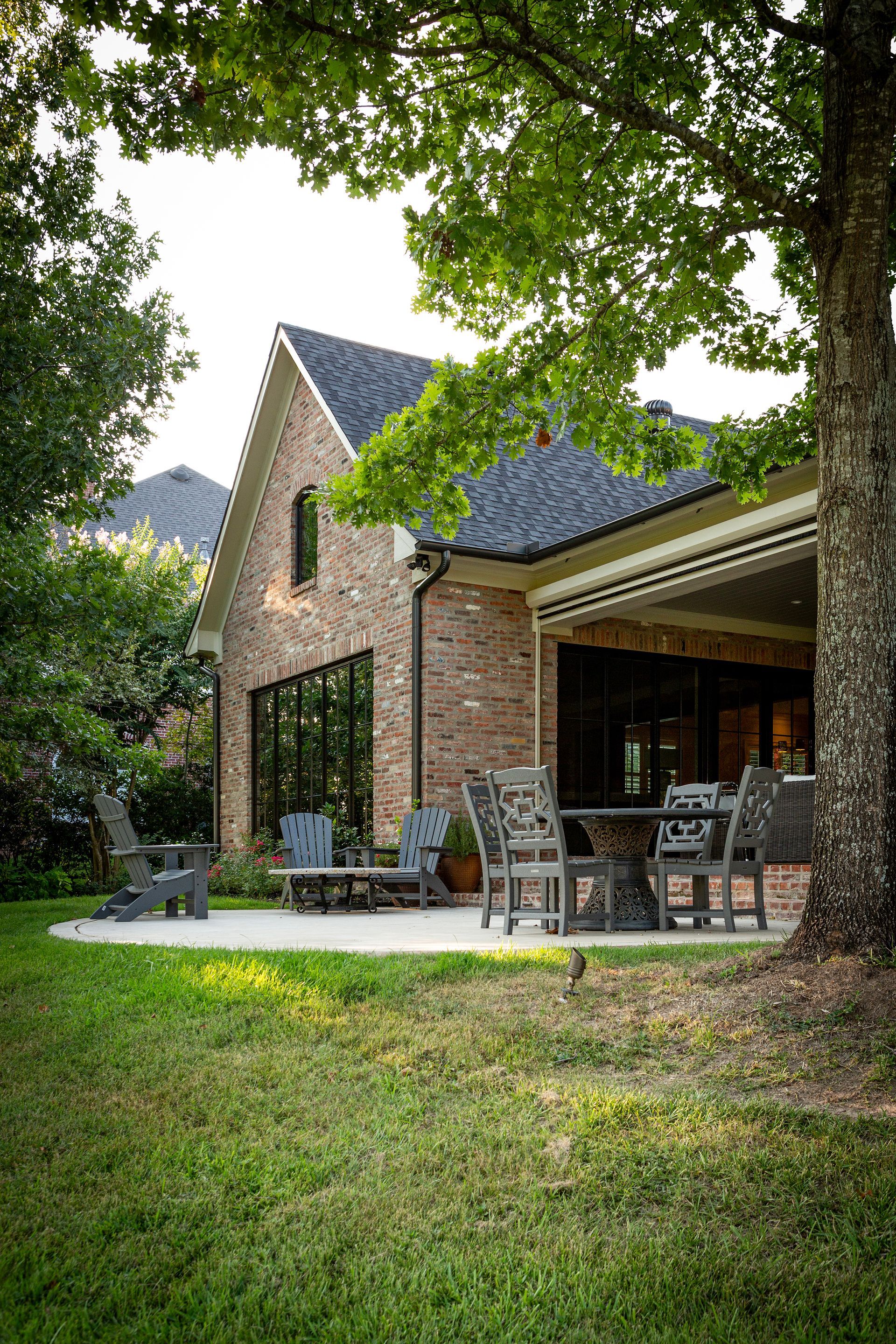 Brick house patio with outdoor chairs under a leafy tree canopy and green lawn