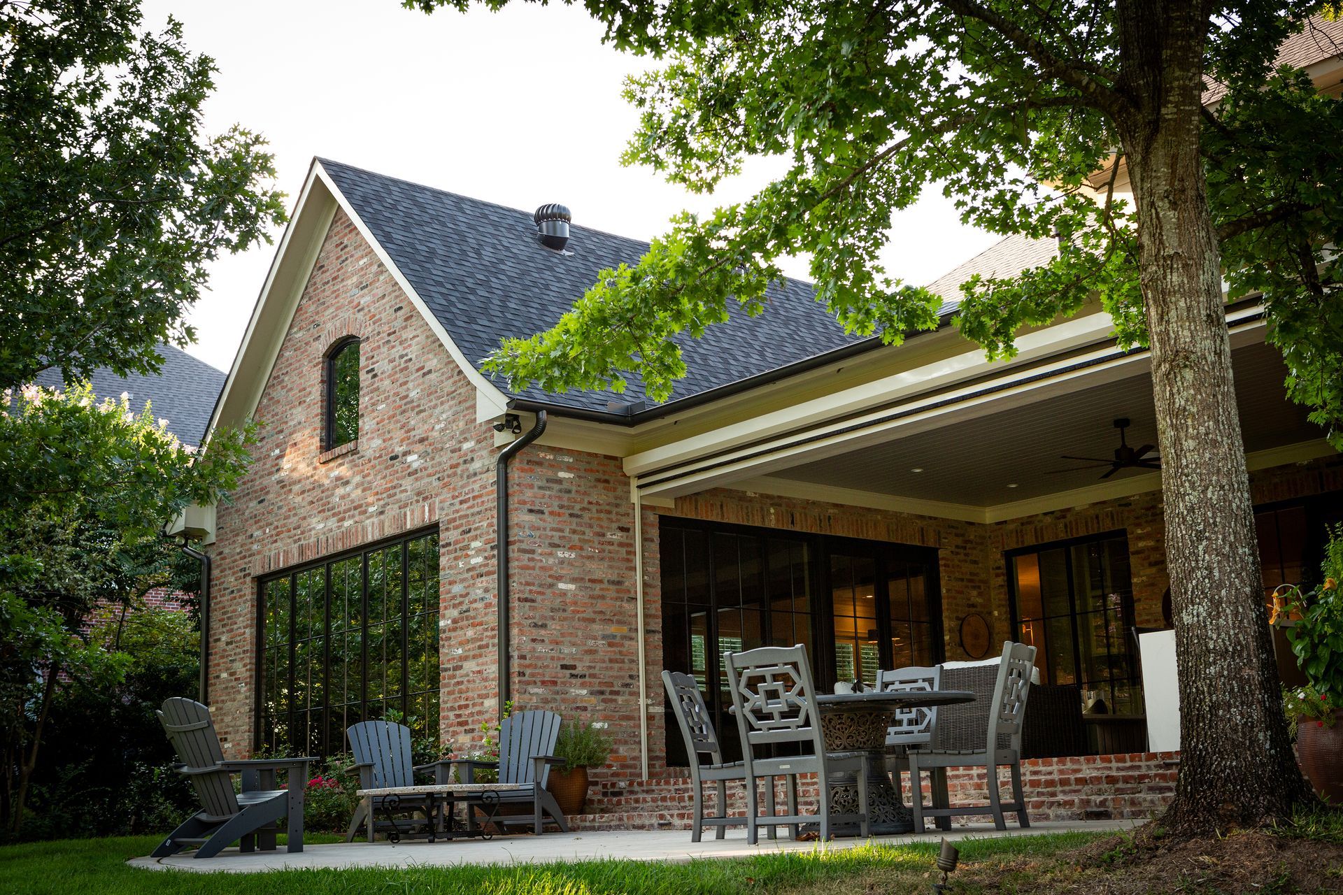 Brick house with covered patio, outdoor dining table, and tall trees in the yard