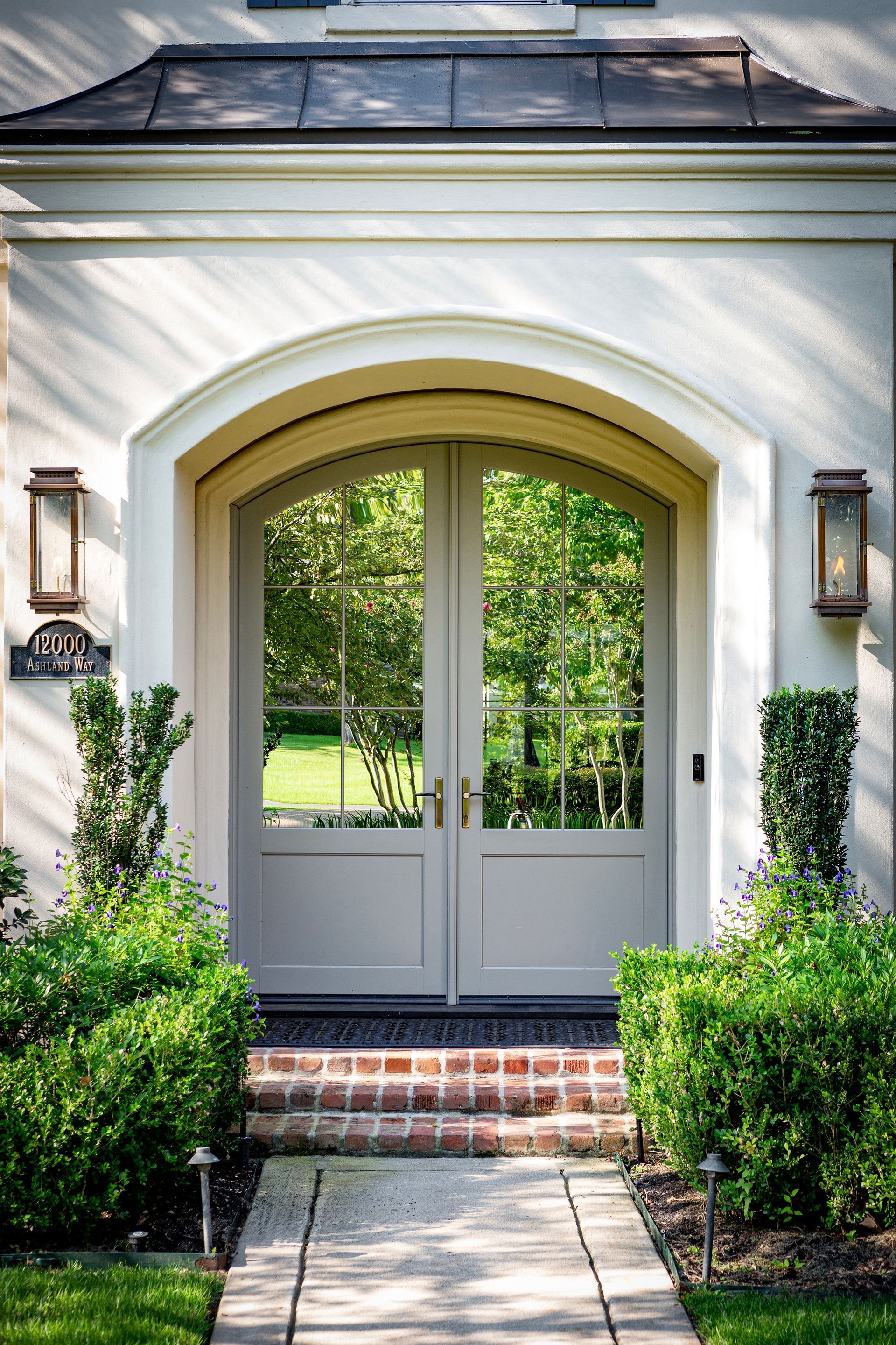 White front entry with arched double doors, brick steps, and trimmed shrubs in a sunny garden.